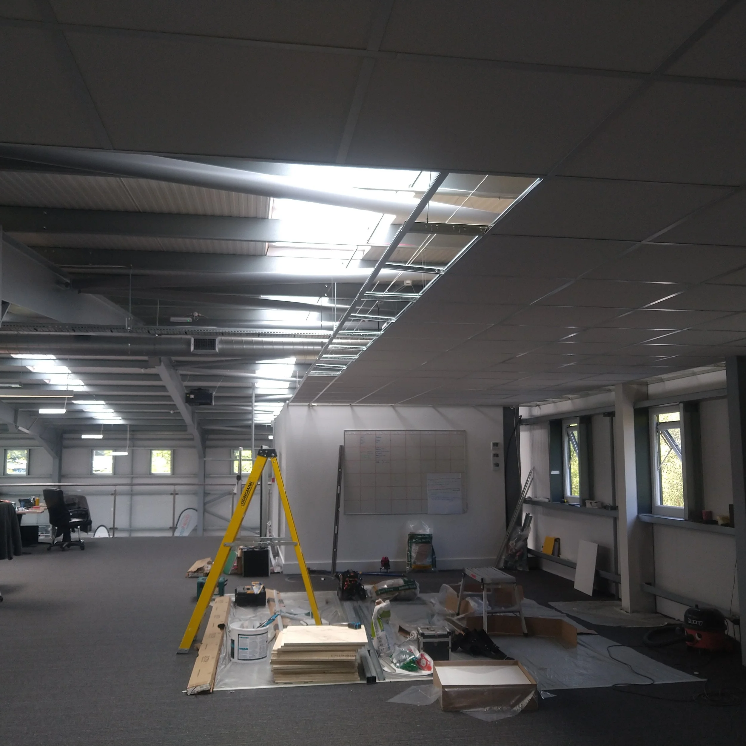 Office space under construction or renovation with tools, materials, and equipment scattered on the floor, a yellow ladder, and a whiteboard on the wall.