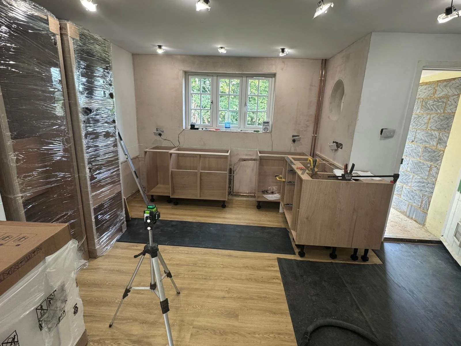Kitchen under renovation with wooden cabinets, some on wheels, and a window. Construction tools and equipment are present, including a tripod and packaging.