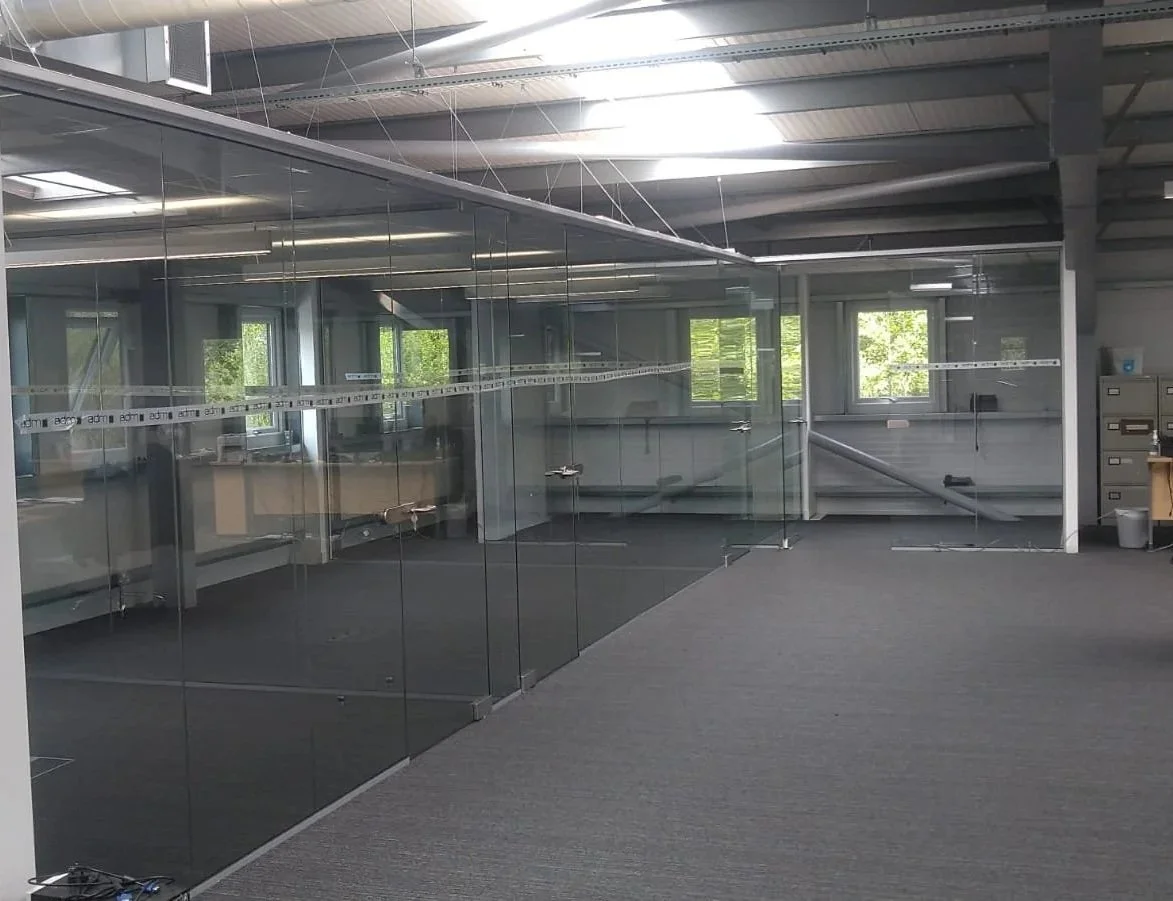 Empty office space with glass walls and gray carpet, with overhead lighting and some windows showing greenery outside.