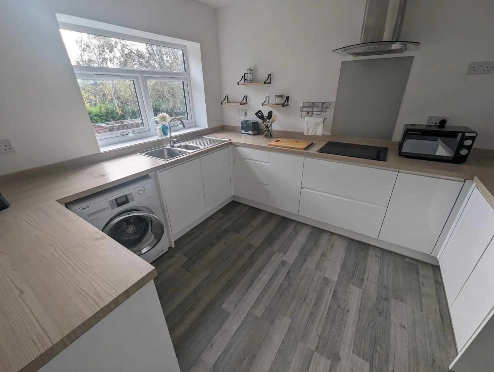 Modern kitchen with white cabinets, wooden countertops, a window above the sink, and kitchen appliances including a washing machine, microwaves, and a cooktop.