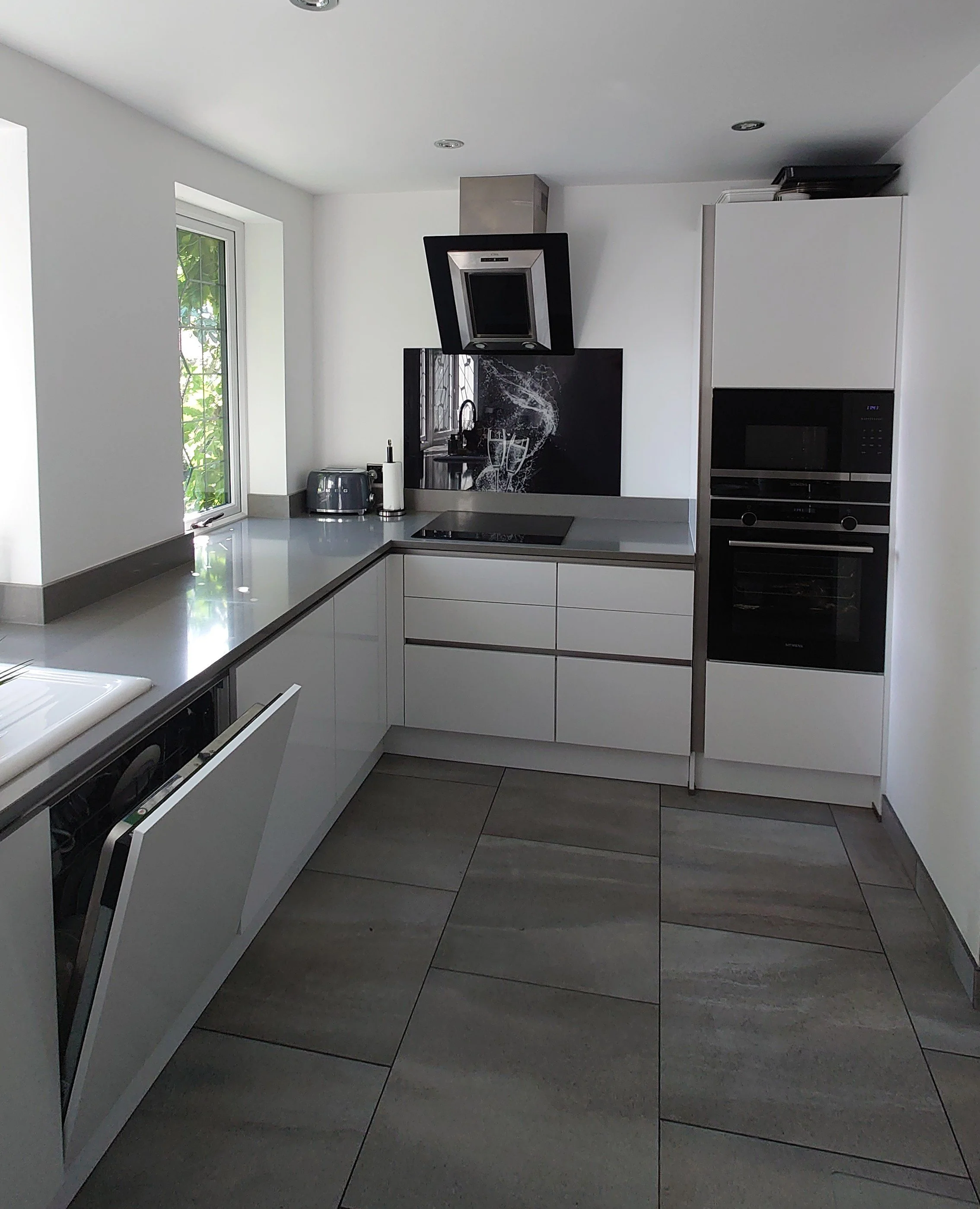 Modern kitchen with white cabinets, gray countertop, black appliances, a window with greenery outside, and a partial view of the floor tiles.