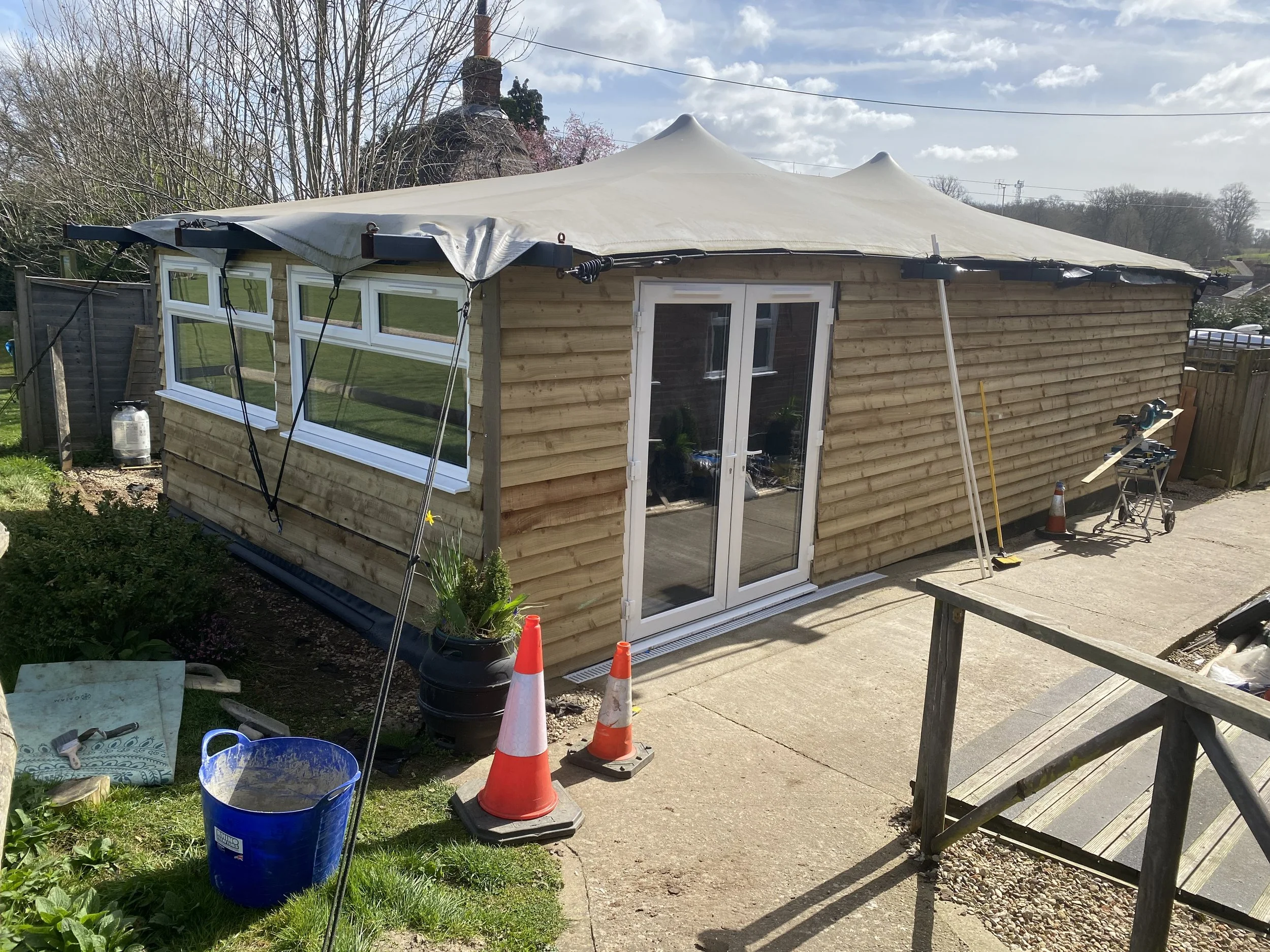 A backyard shed with wooden siding, large windows, sliding glass doors, and a canopy on a sunny day. Construction tools, orange traffic cones, and gardening items are nearby, with a garden area and a wooden deck in the surroundings.