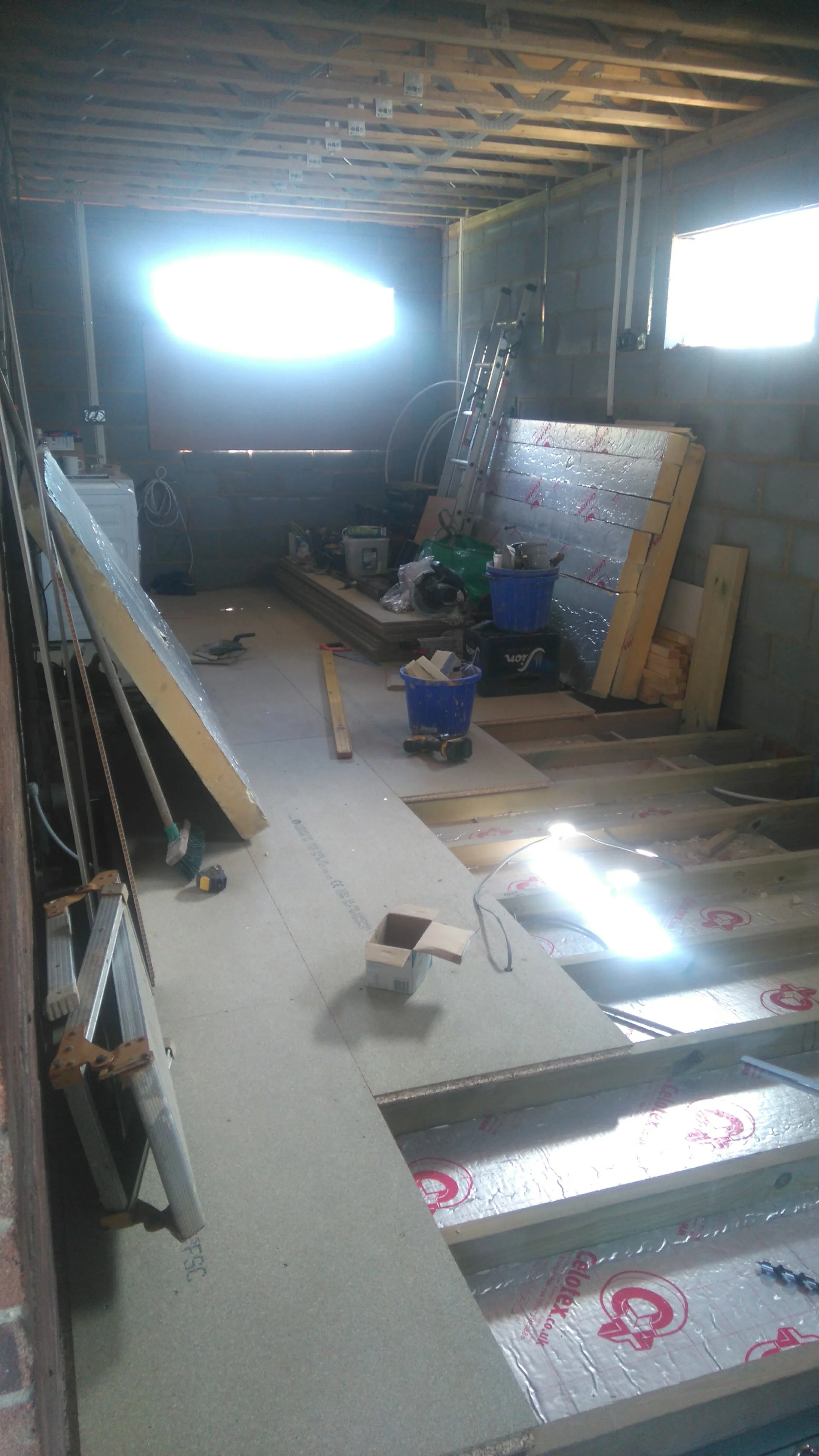 A room under construction with exposed wooden ceiling beams, a cinder block wall, and insulation on the floor. Construction tools, ladders, and materials are scattered around, including buckets, a level, and wood planks.