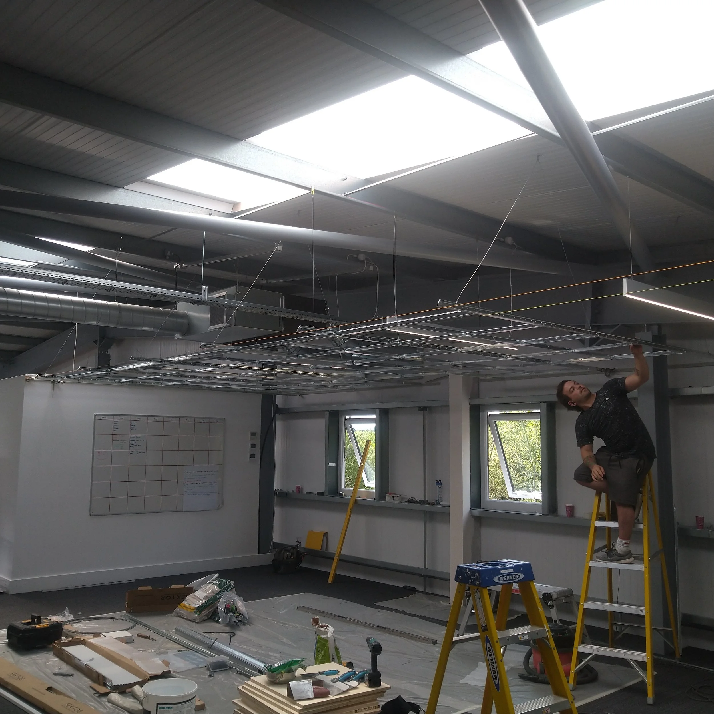 A person is standing on a yellow ladder, working on a suspended ceiling grid in an office under construction, with tools and materials on the floor.