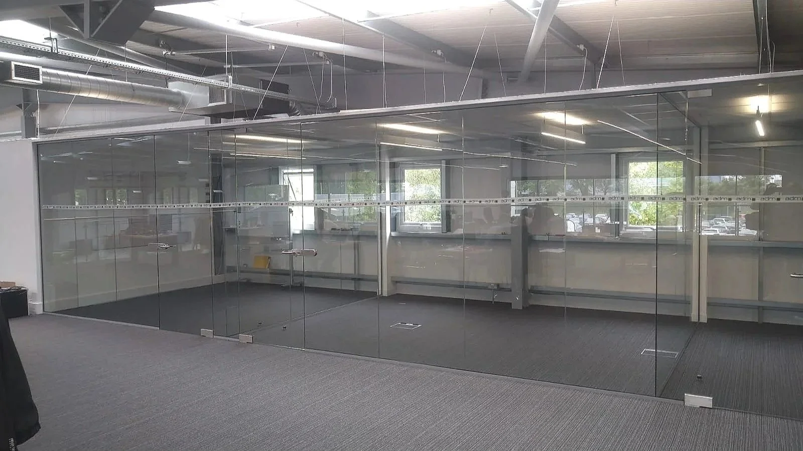 Empty office room with glass walls, ceiling pipes, and large windows showing parked cars outside.