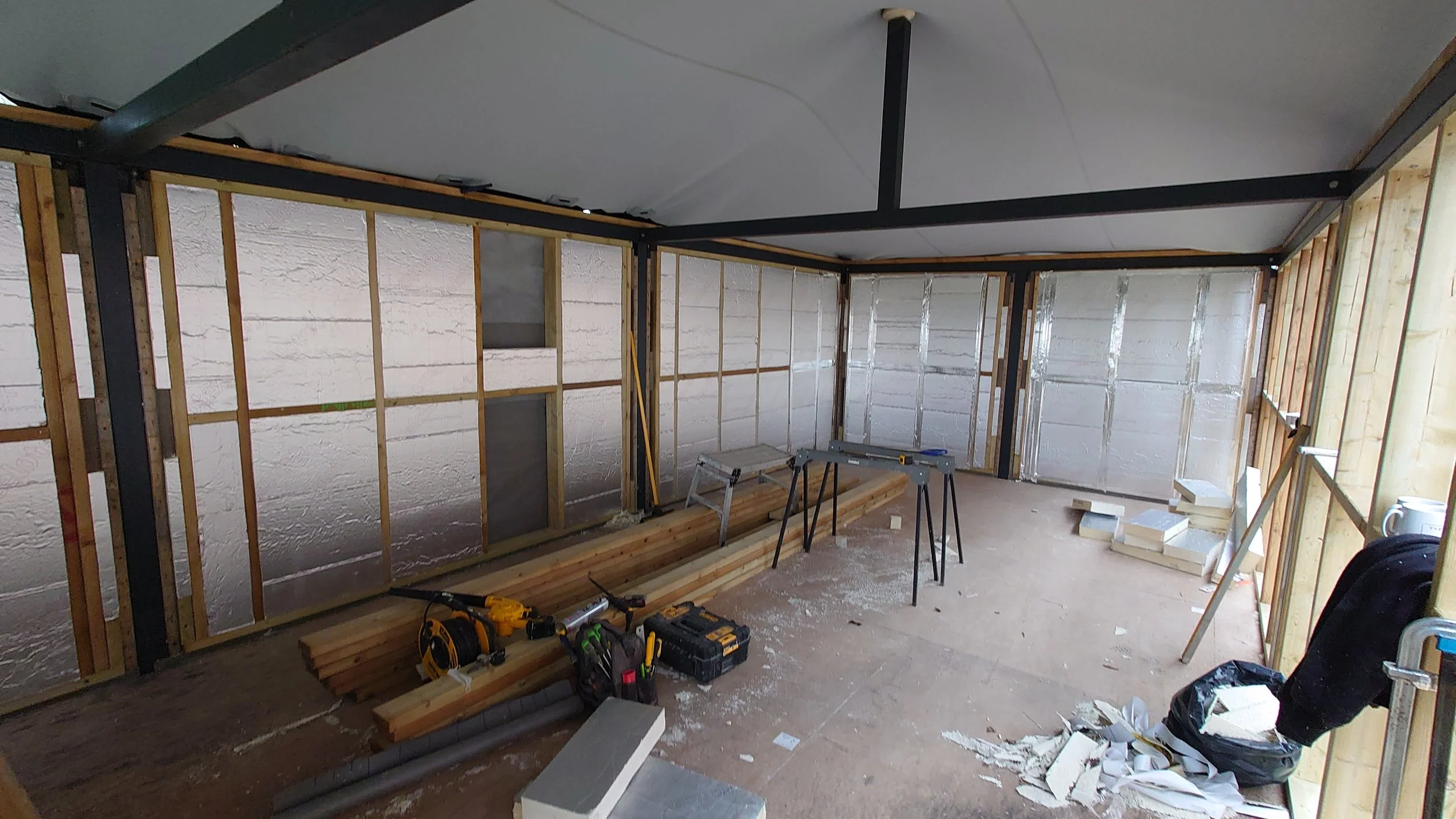 Room under construction with exposed wall framing, insulation, and construction tools including a miter saw, toolbox, screwdriver, and wood planks.