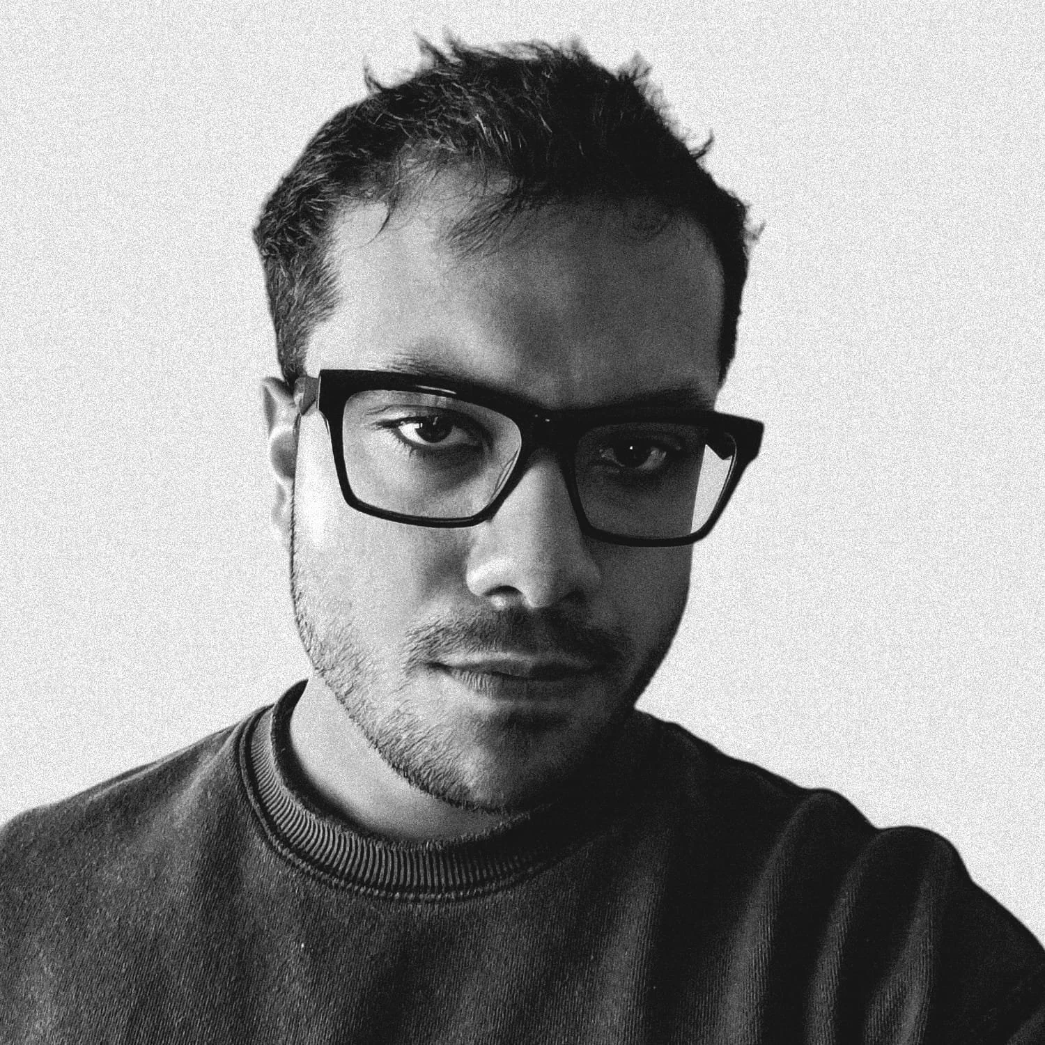 A black-and-white portrait of a young man wearing glasses and a dark sweater, looking directly at the camera.
