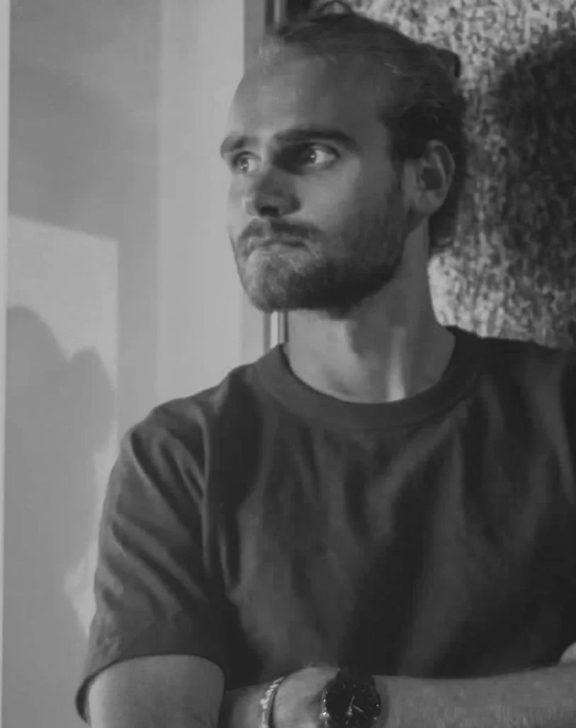 A black-and-white photo of a man with a beard and styled hair, looking to the side. He is wearing a dark T-shirt and a wristwatch, with arms crossed. The background features a textured wall.