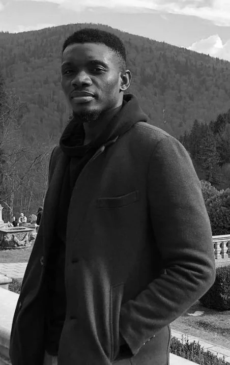 A young man in a black coat standing outdoors with hills and trees in the background.