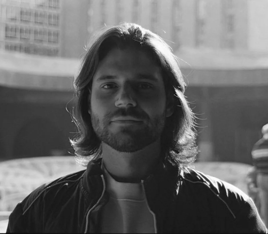 A young man with shoulder-length hair, a beard, and mustache, standing outdoors with a cityscape in the background, in black and white.