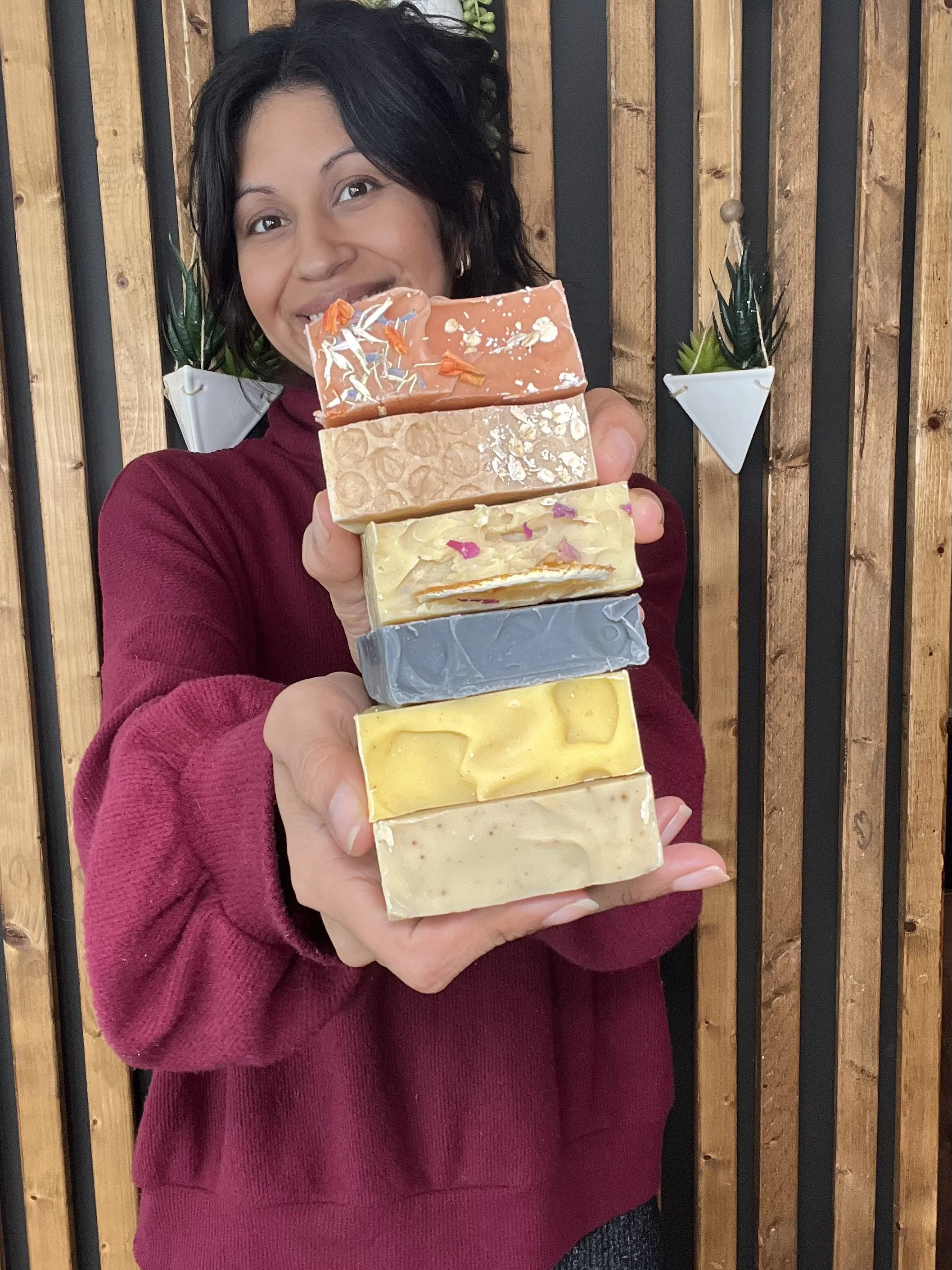 Woman with dark hair smiling and holding a stack of six colorful handmade soap bars in front of a wooden wall with decorative plants.