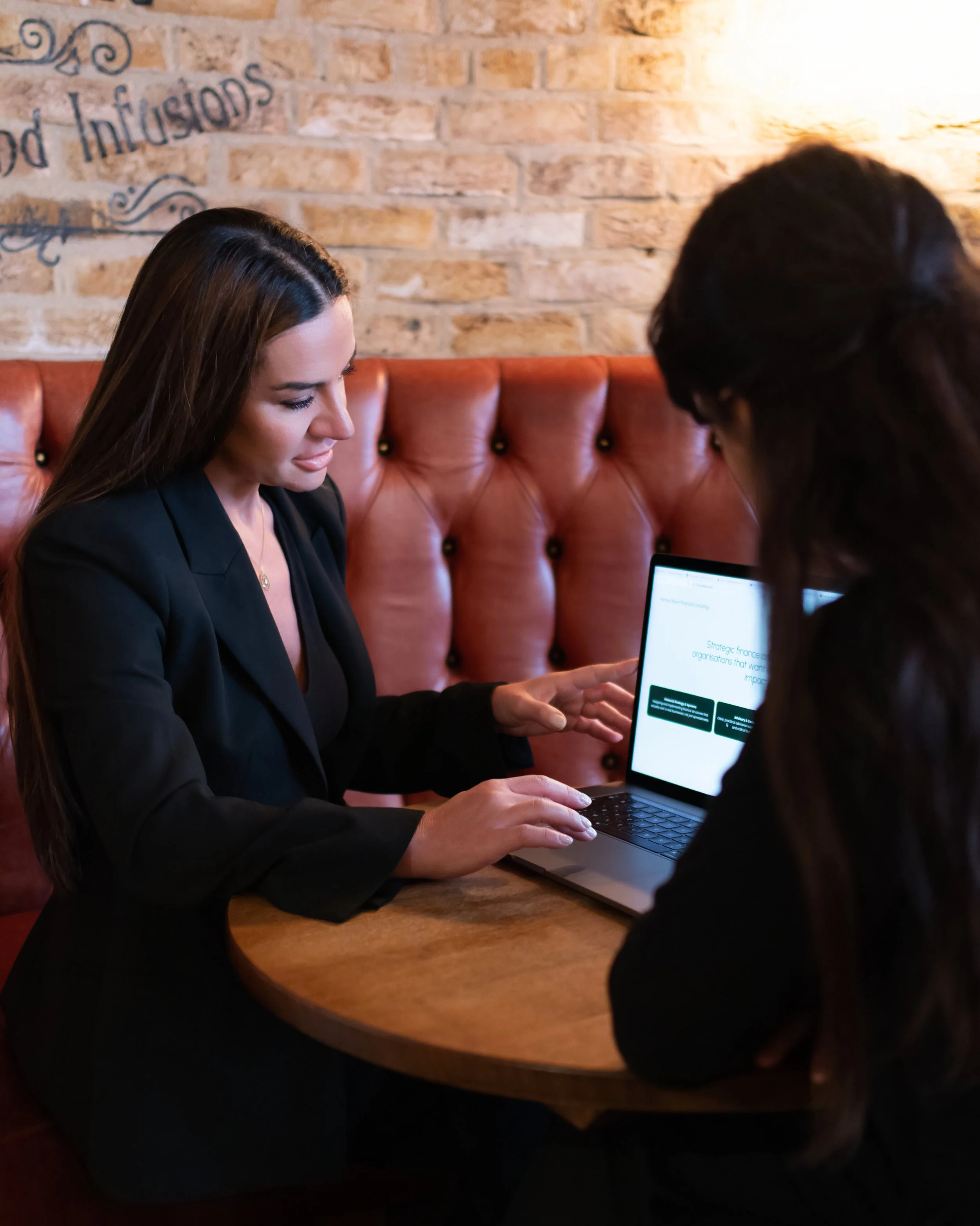 Two women in business attire sitting at a round wooden table in a cafe, looking at a laptop screen with a brick wall in the background.