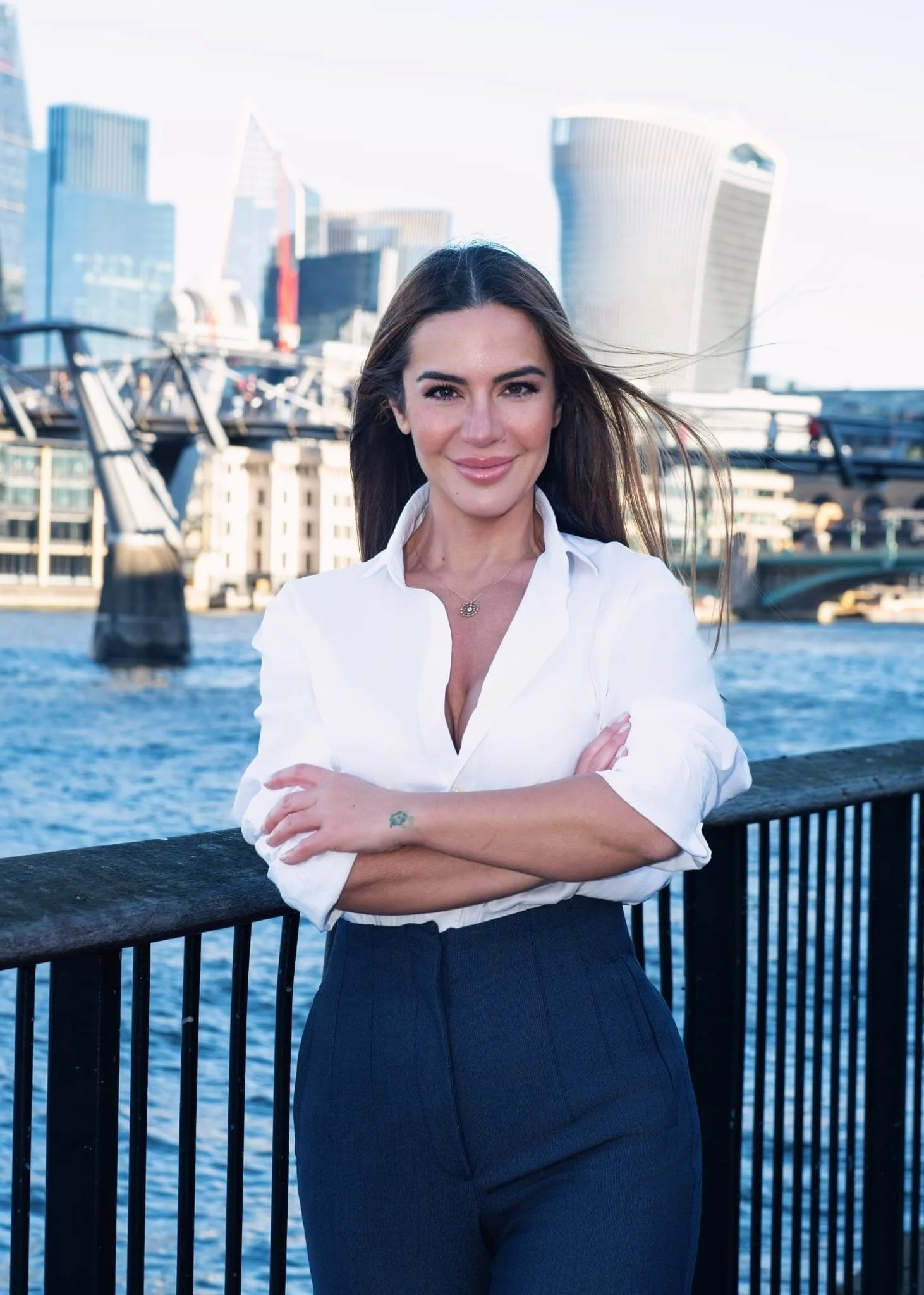A woman with long dark hair, wearing a white blouse and dark pants, stands in front of a body of water with a city skyline in the background. She has her arms crossed and is smiling at the camera.