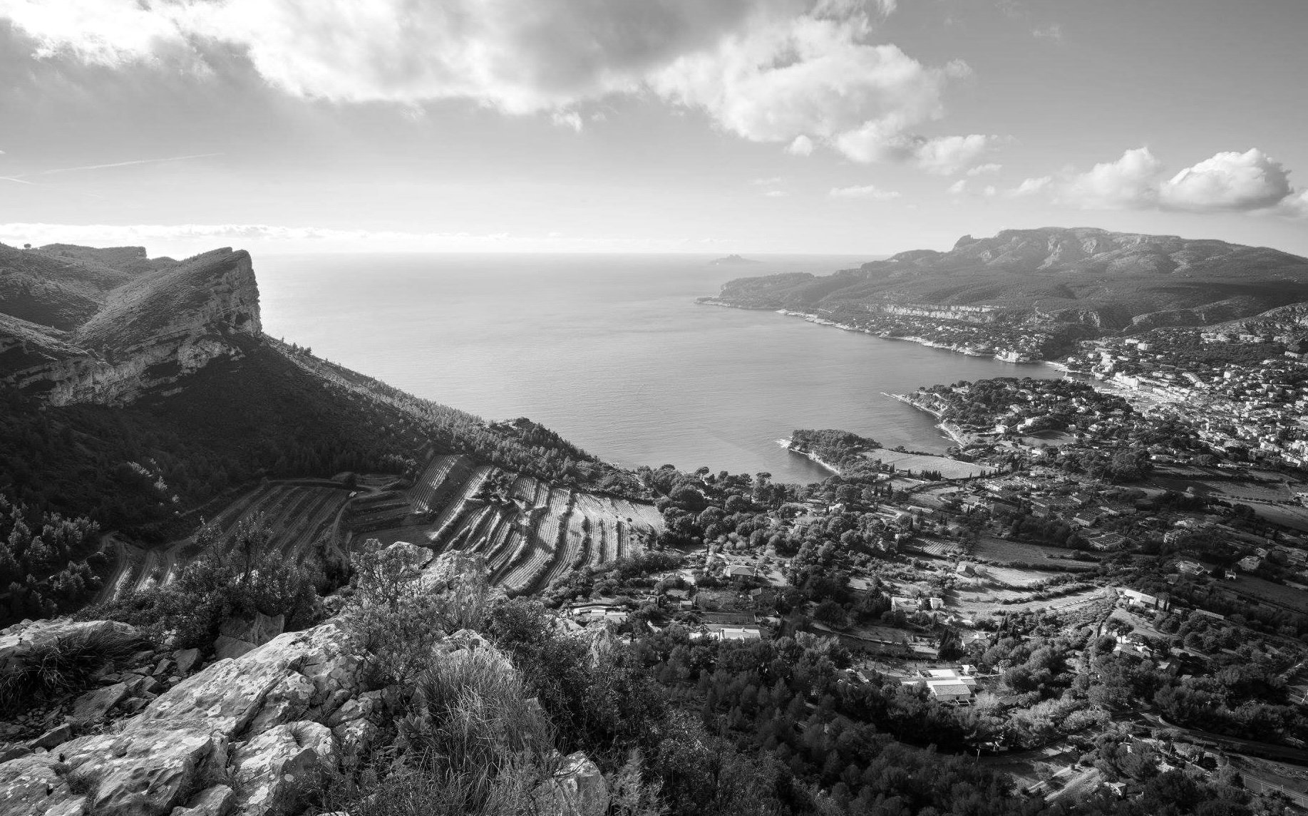 A black and white aerial view of a coastal landscape in Provence, France featuring mountains, a view of the Mediterranean, and the town of Cassis on the shoreline with terraced fields on the hillside.