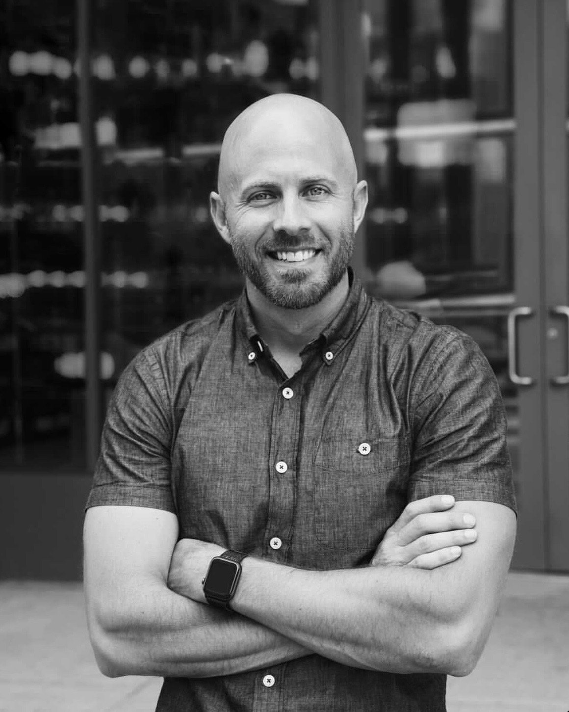 Black and white photo of Dustin Wilson MS, crossed arms, wearing a short-sleeved button-up shirt and a smartwatch, standing outside in front of a glass building.