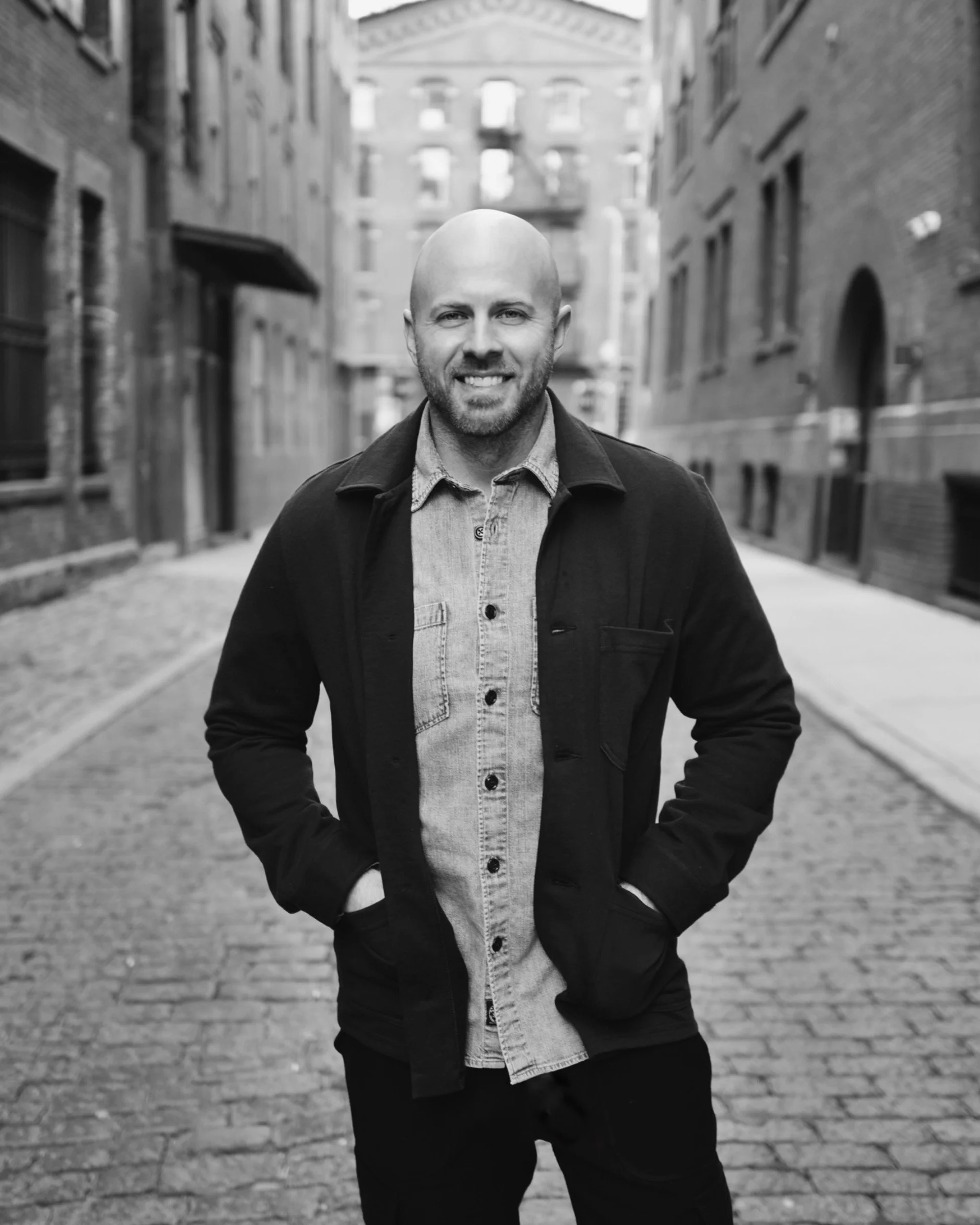 Dustin Wilson MS standing on a cobblestone street in Tribeca, NYC, wearing a light denim shirt and a dark jacket with hands in pockets, with old brick buildings in the background