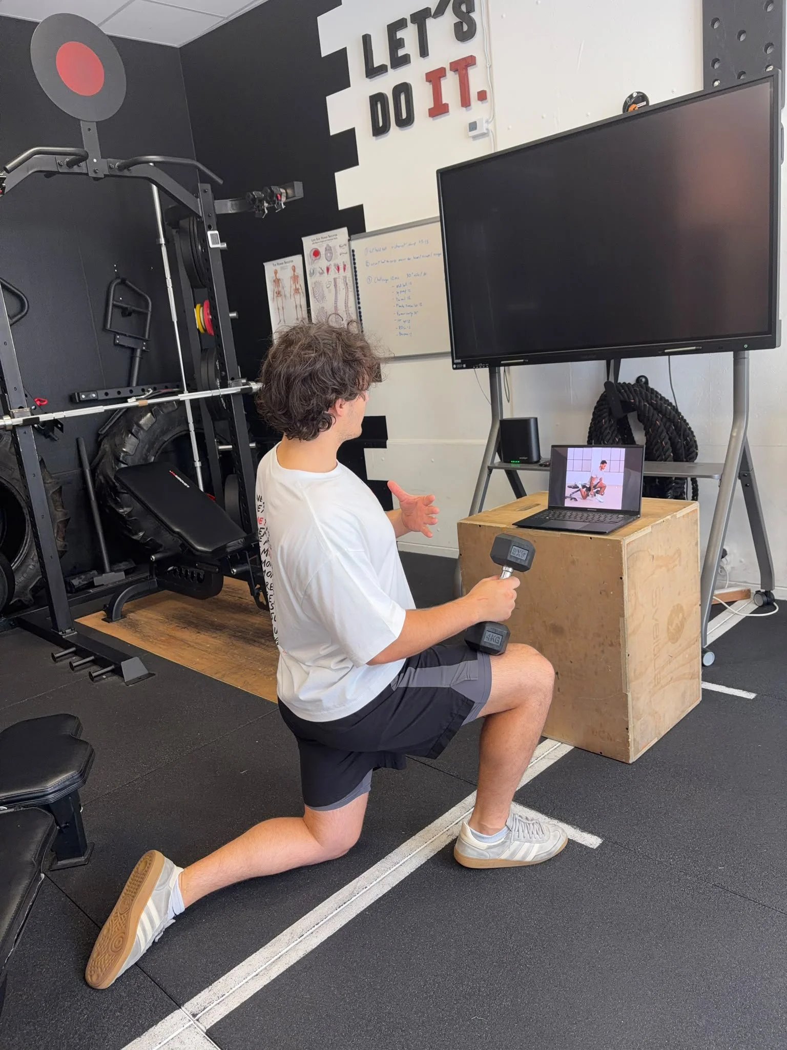 Un homme faisant un exercice de renforcement musculaire dans une salle de sport, en position de fente avant, tenant un haltère dans la main droite, devant un écran de télévision et un tableau blanc.