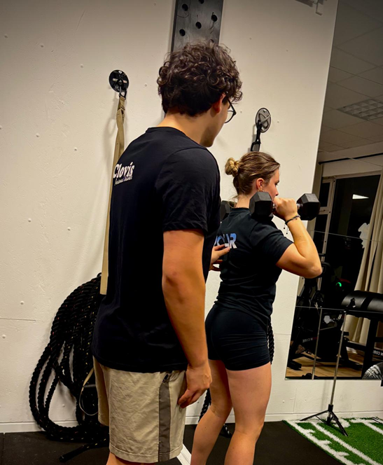 Deux personnes s'entraînent en salle de sport, dont une femme faisant des exercices avec des haltères, vue de dos.