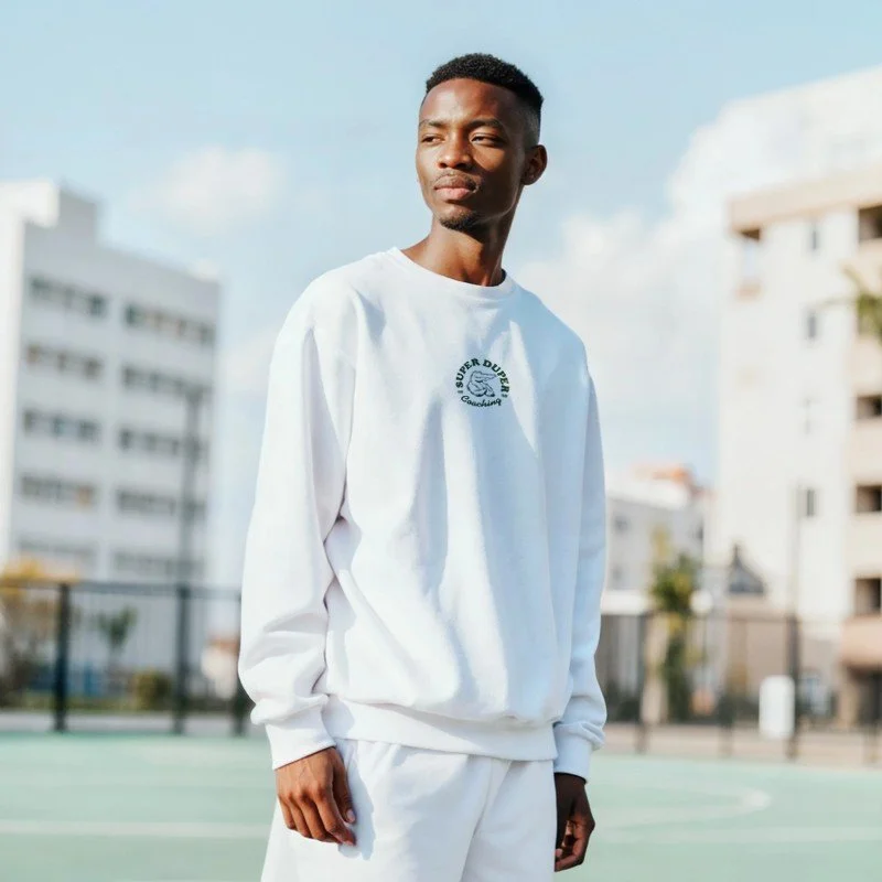 Jeune homme africain debout dans un terrain de sport en plein air, portant un sweat blanc et un pantalon blanc, avec un bâtiment moderne en arrière-plan, sous un ciel bleu avec quelques nuages.