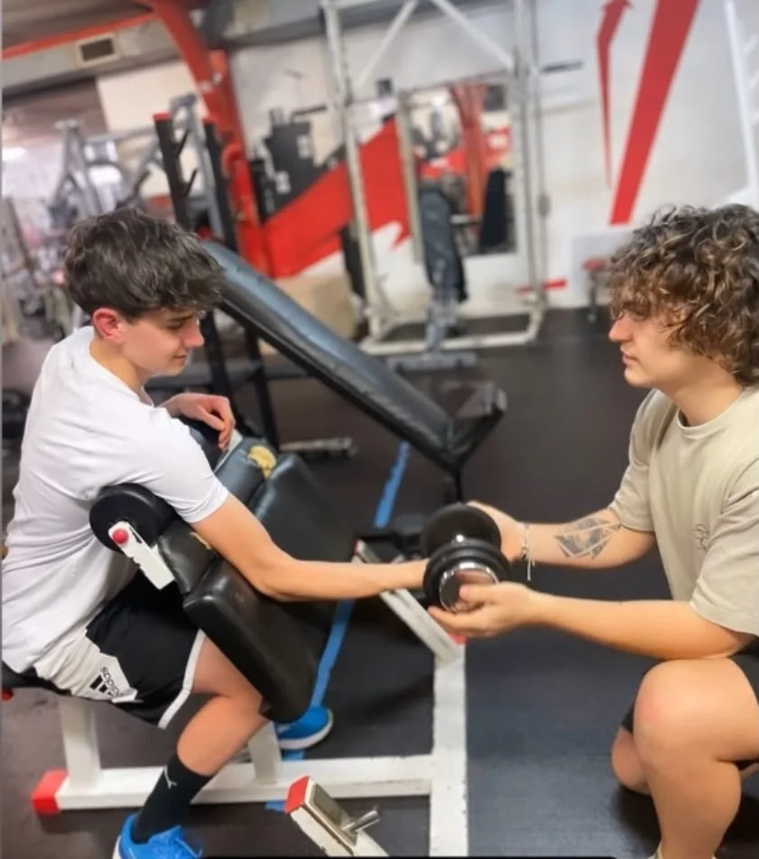 Un adolescent, assis sur un banc de musculation, s'entraîne avec un poids de musculation assisté par une autre personne dans une salle de sport.