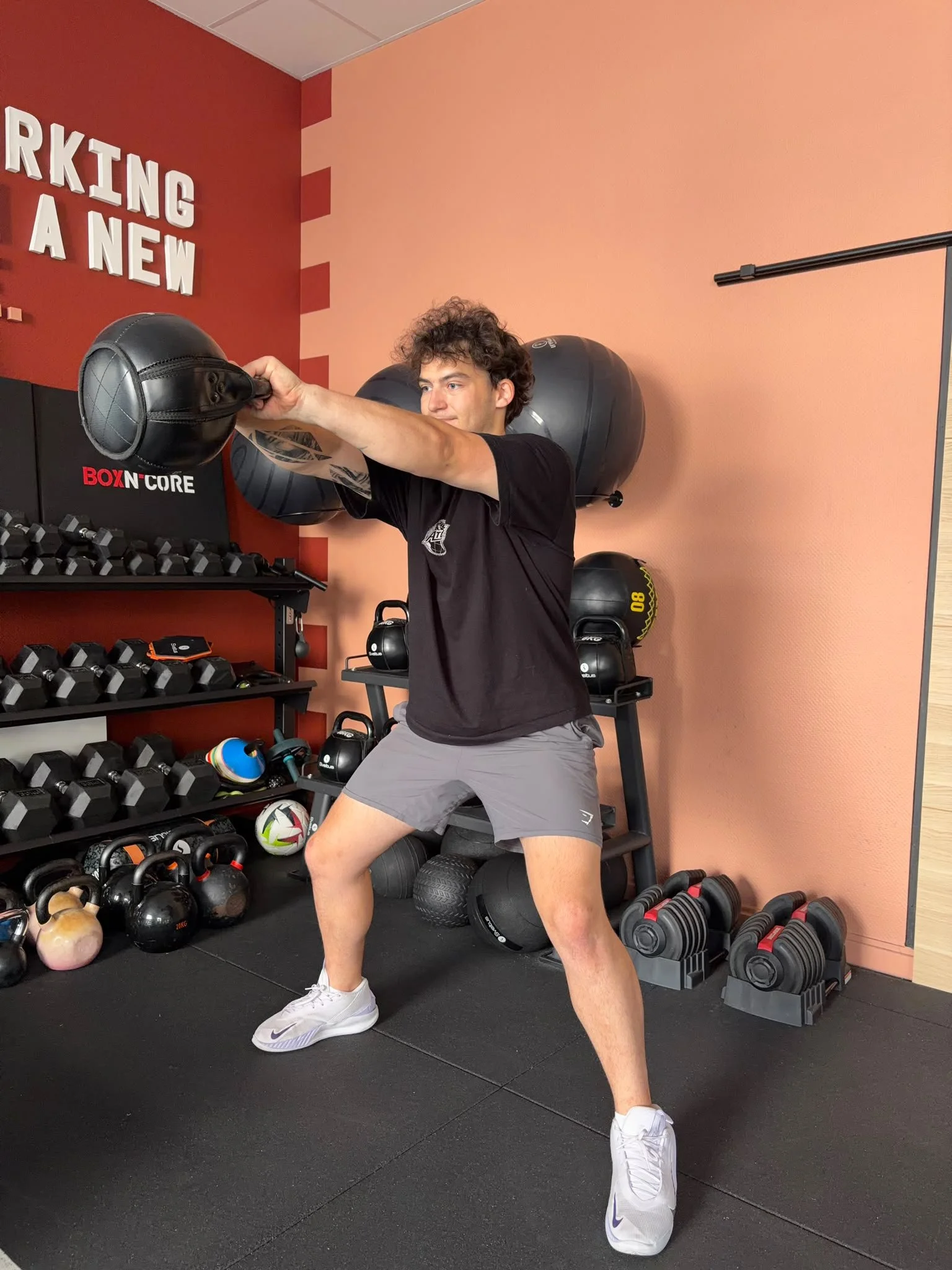 Un homme s'entraîne en soulevant un haltère avec ses bras, dans une salle de sport. Il porte un t-shirt noir, des shorts gris et des chaussures blanches. Derrière lui, il y a des équipements de musculation, des balles de sport, et un mur peint en rose pâle et rouge avec une phrase