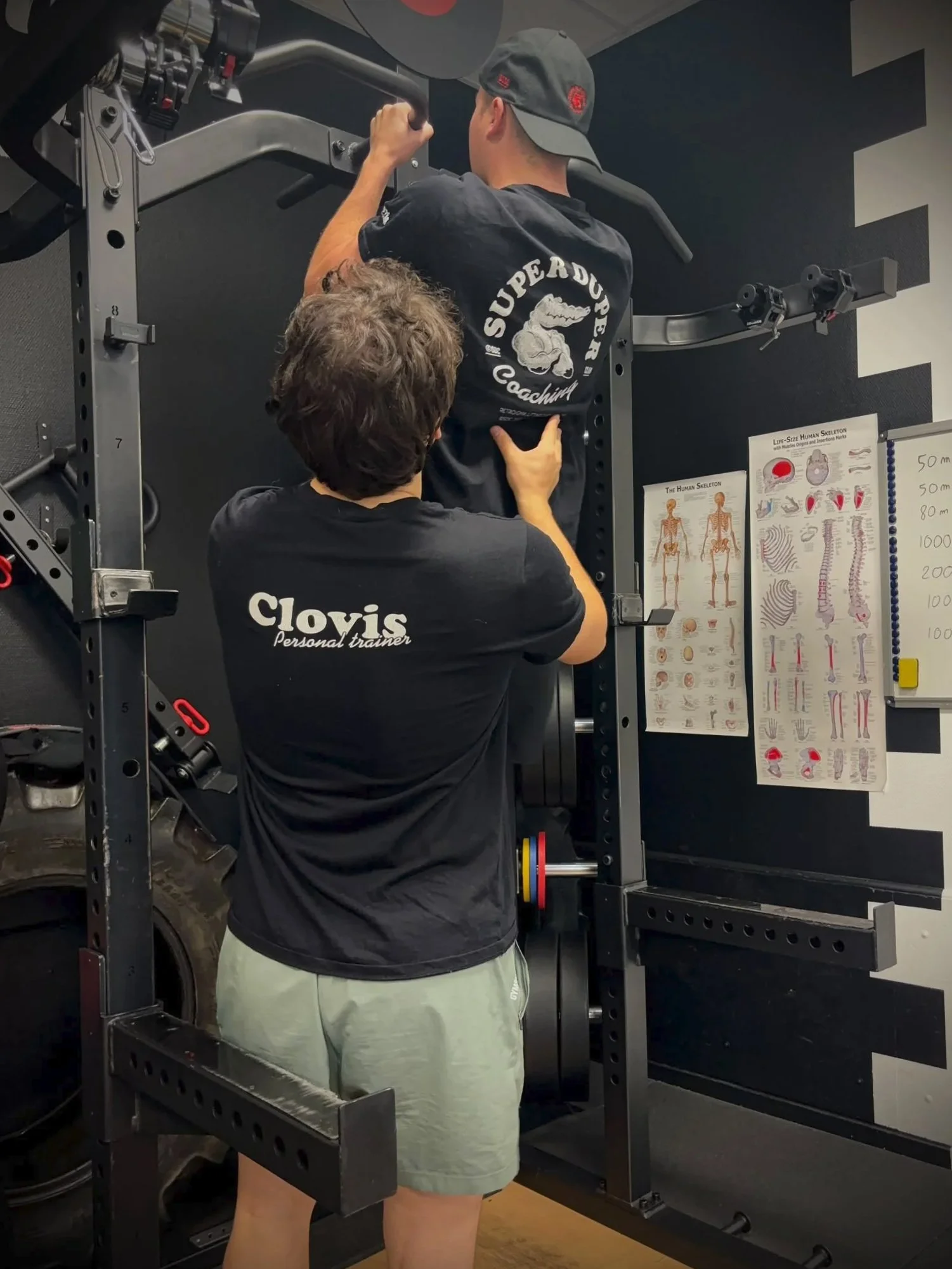 Un entraîneur personnel aide un homme à faire un exercice de musculation sur une machine de musculation en salle de sport. La salle a des posters anatomiques sur le mur.