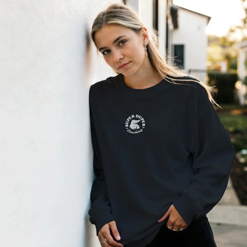 Jeune femme qui se cache contre un mur blanc, portant un sweat noir avec un logo blanc » SUPER DUFFER Coaching » à l'avant, en extérieur dans une rue pavée, soirée ensoleillée.