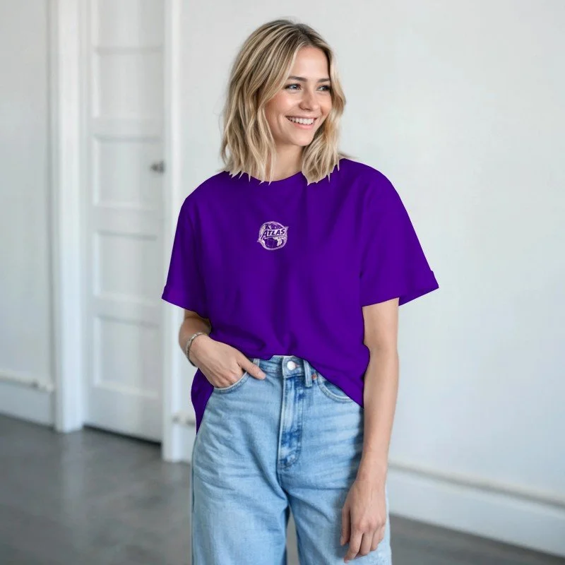 Une femme souriante avec les cheveux blonds, portant un t-shirt violet avec un logo et un jean clair, debout dans une pièce lumineuse avec un mur blanc et une porte en arrière-plan.