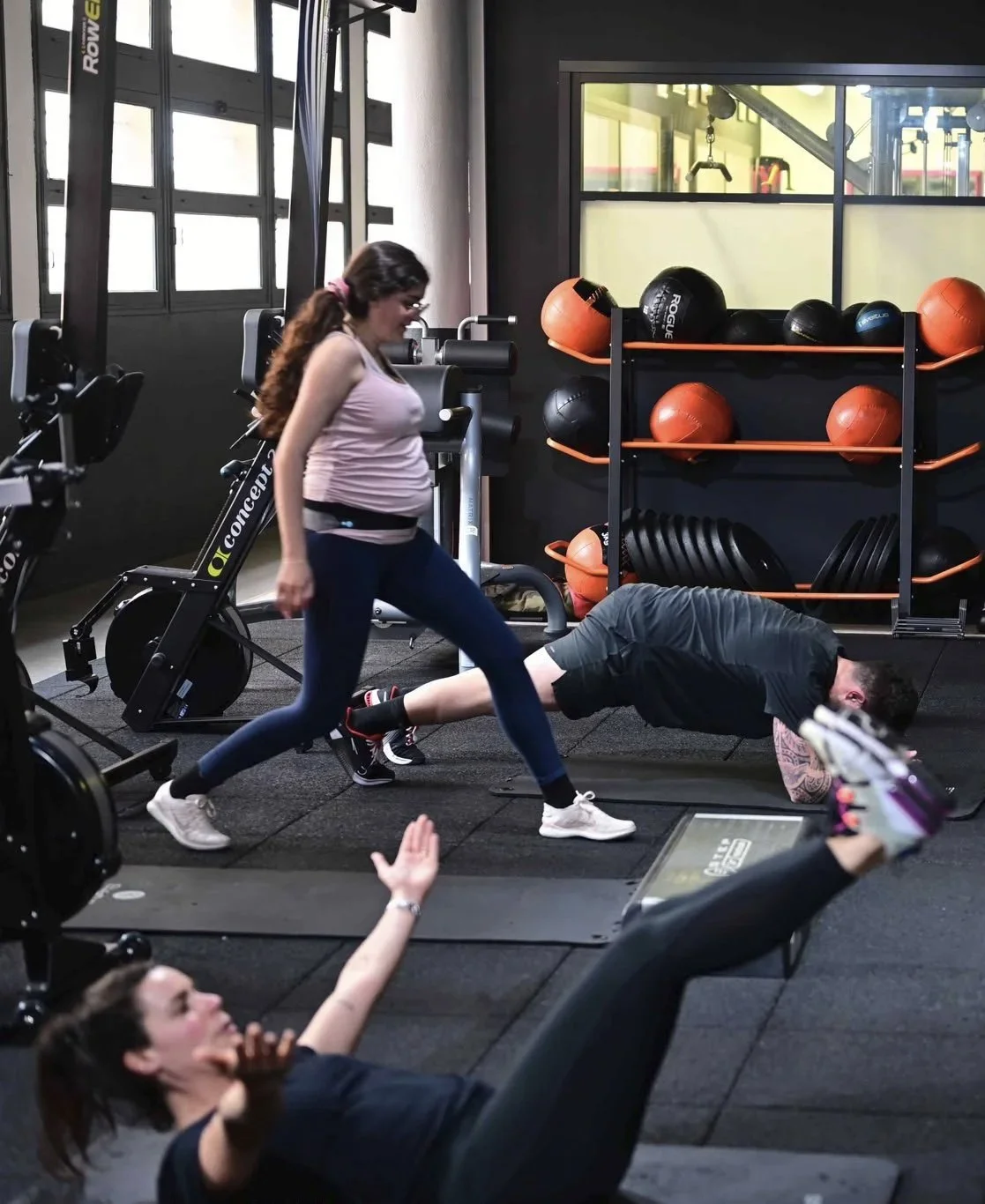 Une personne se fait entraîner par une autre lors d'un entraînement de fitness dans une salle de sport, avec des ballons médicinaux et équipement de musculation en arrière-plan.