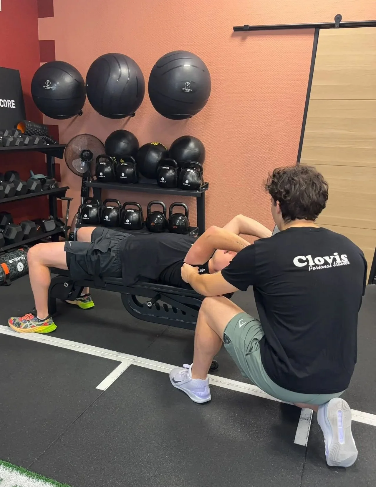 Un entraîneur aide un jeune athlète à faire un étirement ou un exercice de renforcement sur un lit d'entraînement dans une salle de sport. Il porte un t-shirt noir avec le nom 'Clovis Personal Trainer'.
