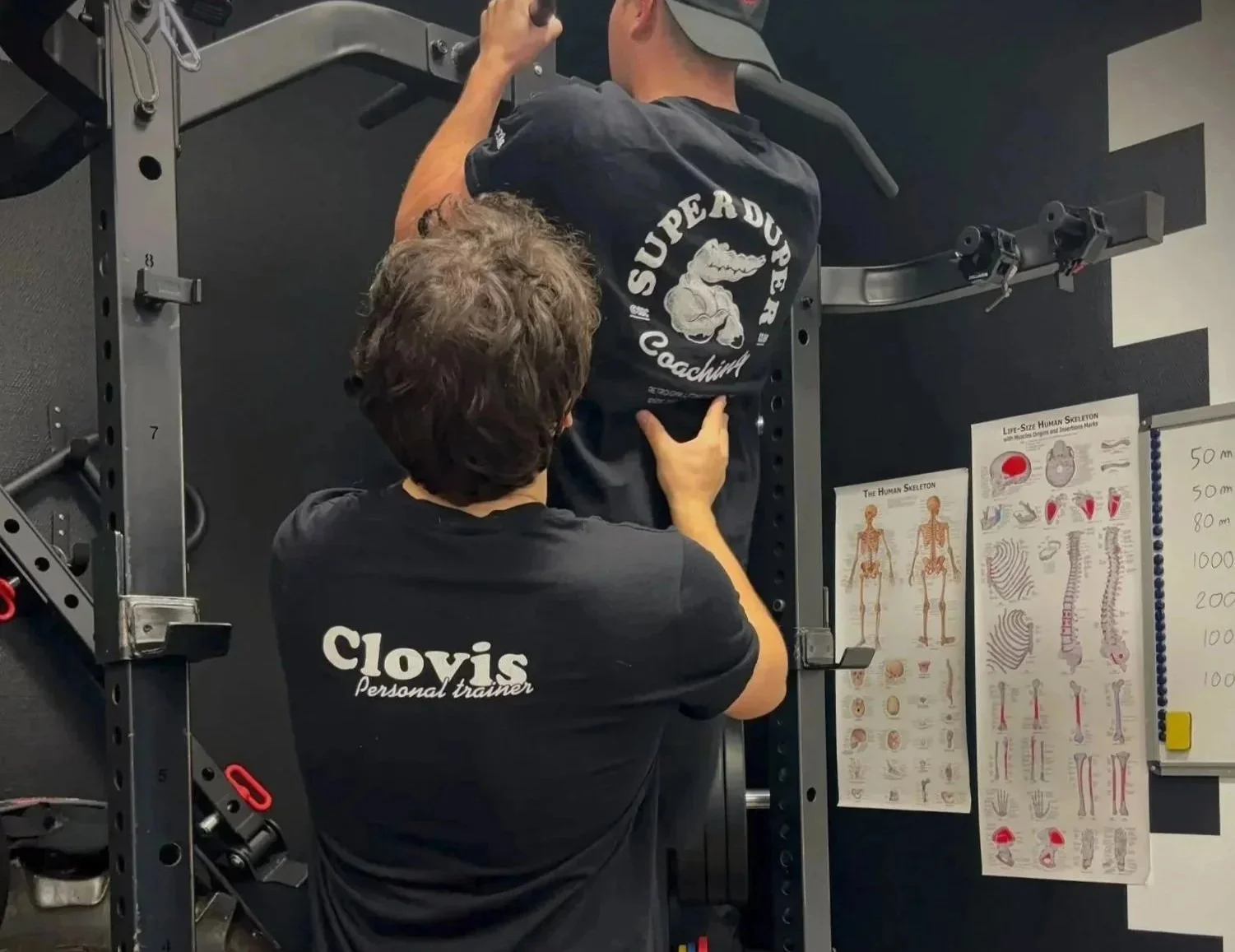 Un entraîneur personnel aidant un homme à faire un exercice de musculation dans une salle de sport, avec des posters anatomiques visibles au mur.