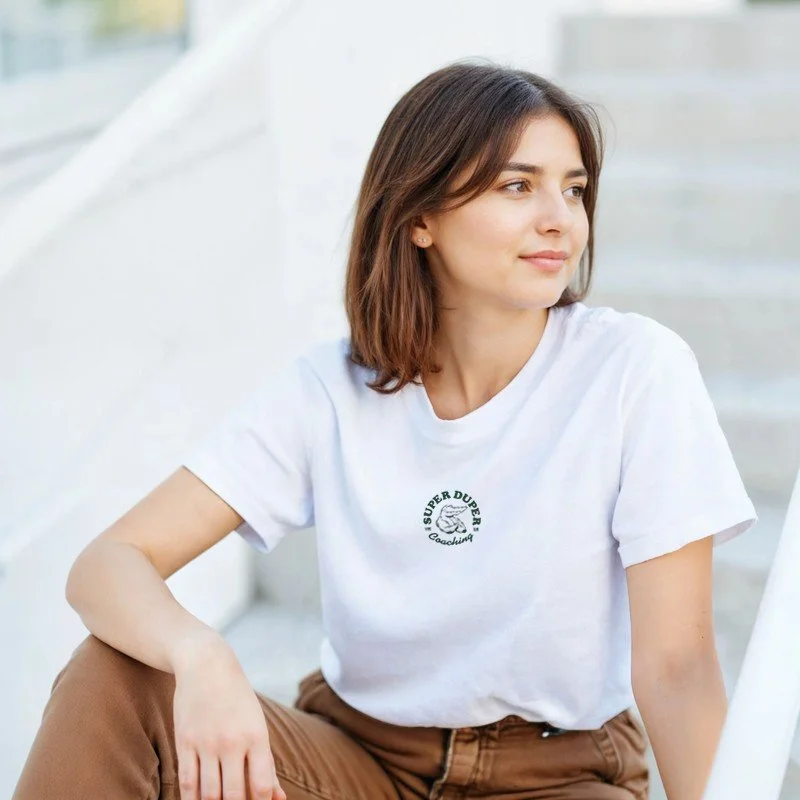 Jeune femme assise à l'extérieur, portant un t-shirt blanc avec un logo et un pantalon marron.