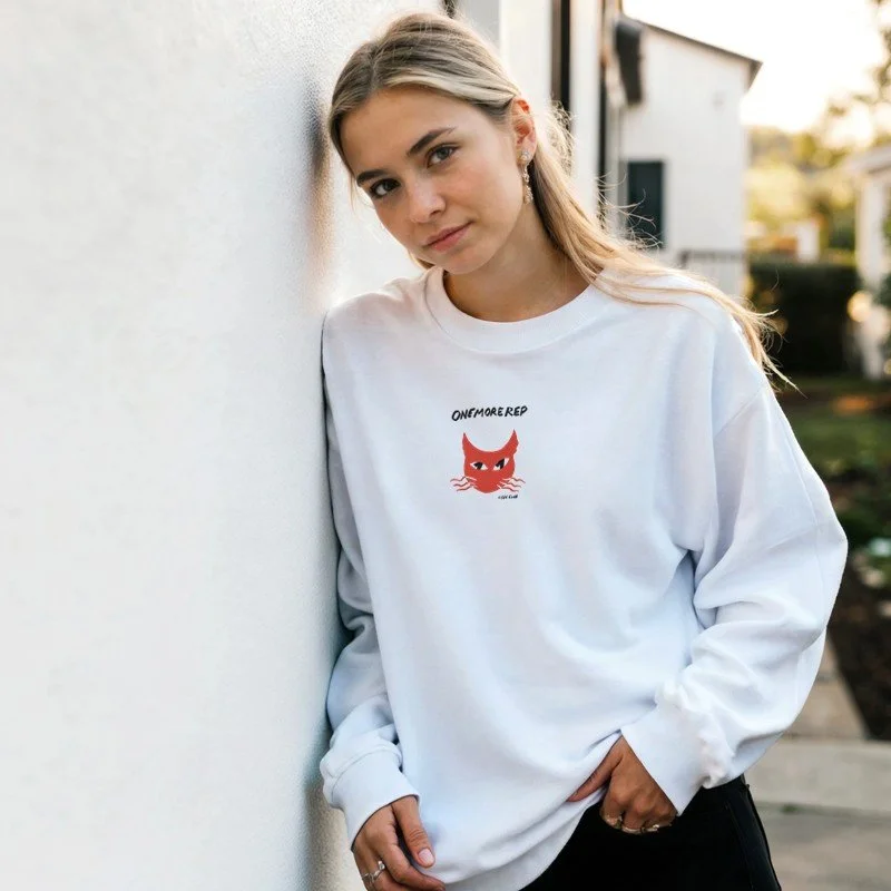 Jeune femme avec des cheveux longs, portant un sweat blanc avec un dessin de chat rouge et la phrase "ONEMORE REP".