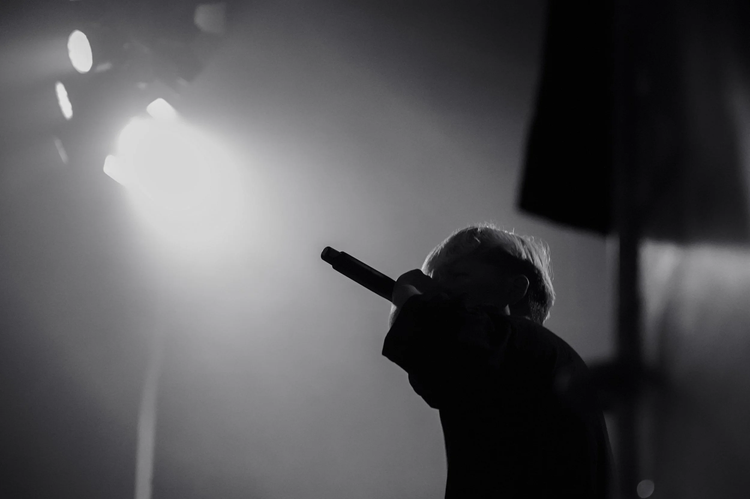 A black and white photo of a person with short hair holding a microphone, facing away from the camera, with stage lights in the background.