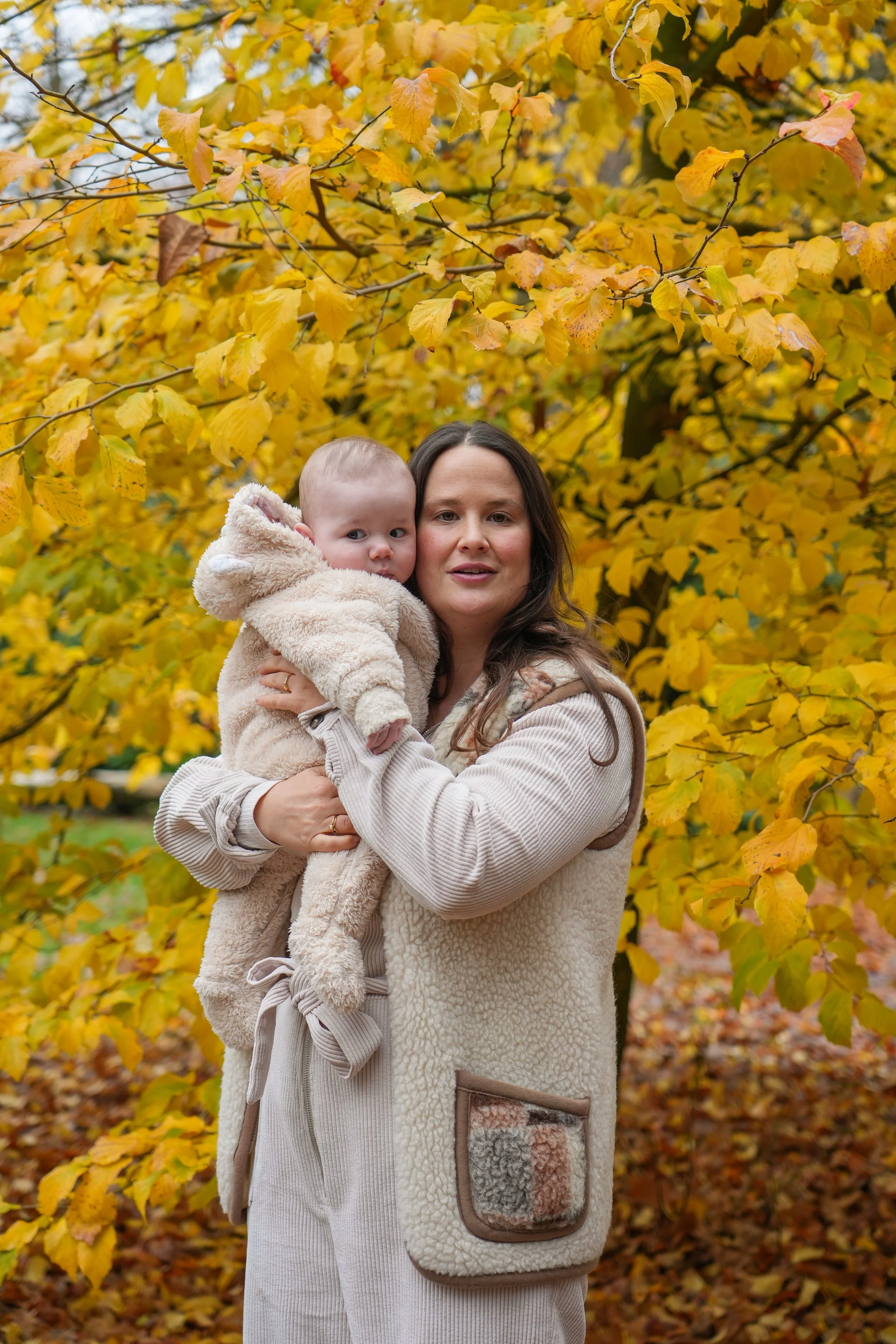 A woman holding a baby in front of a tree with vibrant yellow autumn leaves.