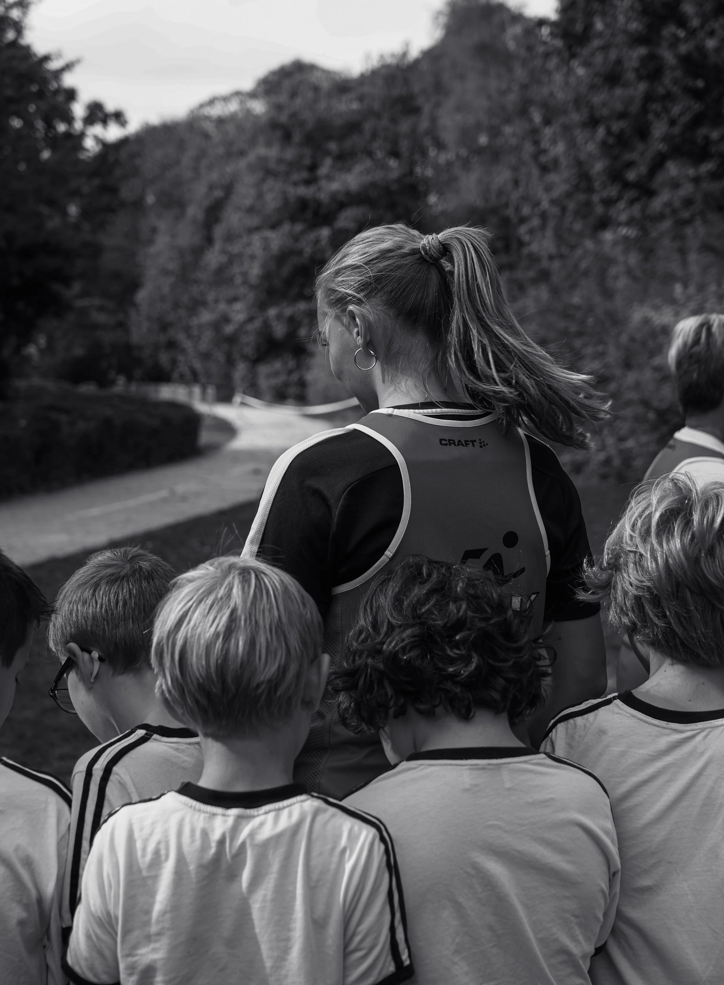 A group of young children gathered outdoors, facing away from the camera, with a woman in a sports jersey with a ponytail and earrings standing among them, possibly listening or waiting for instructions, with trees and a pathway in the background.