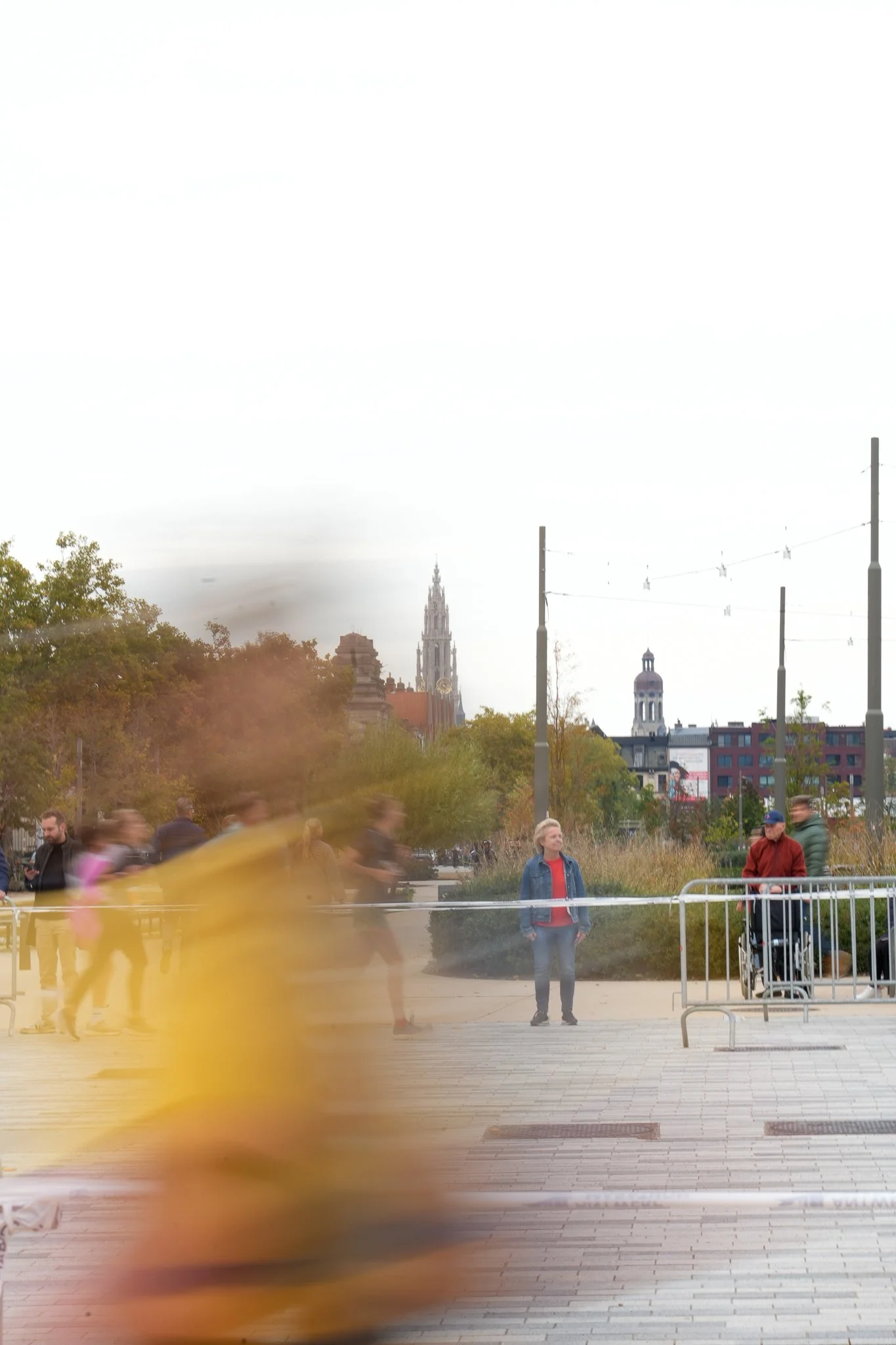 City scene with blurred pedestrians and a woman standing still near railings, with historic buildings and a church tower in the background.