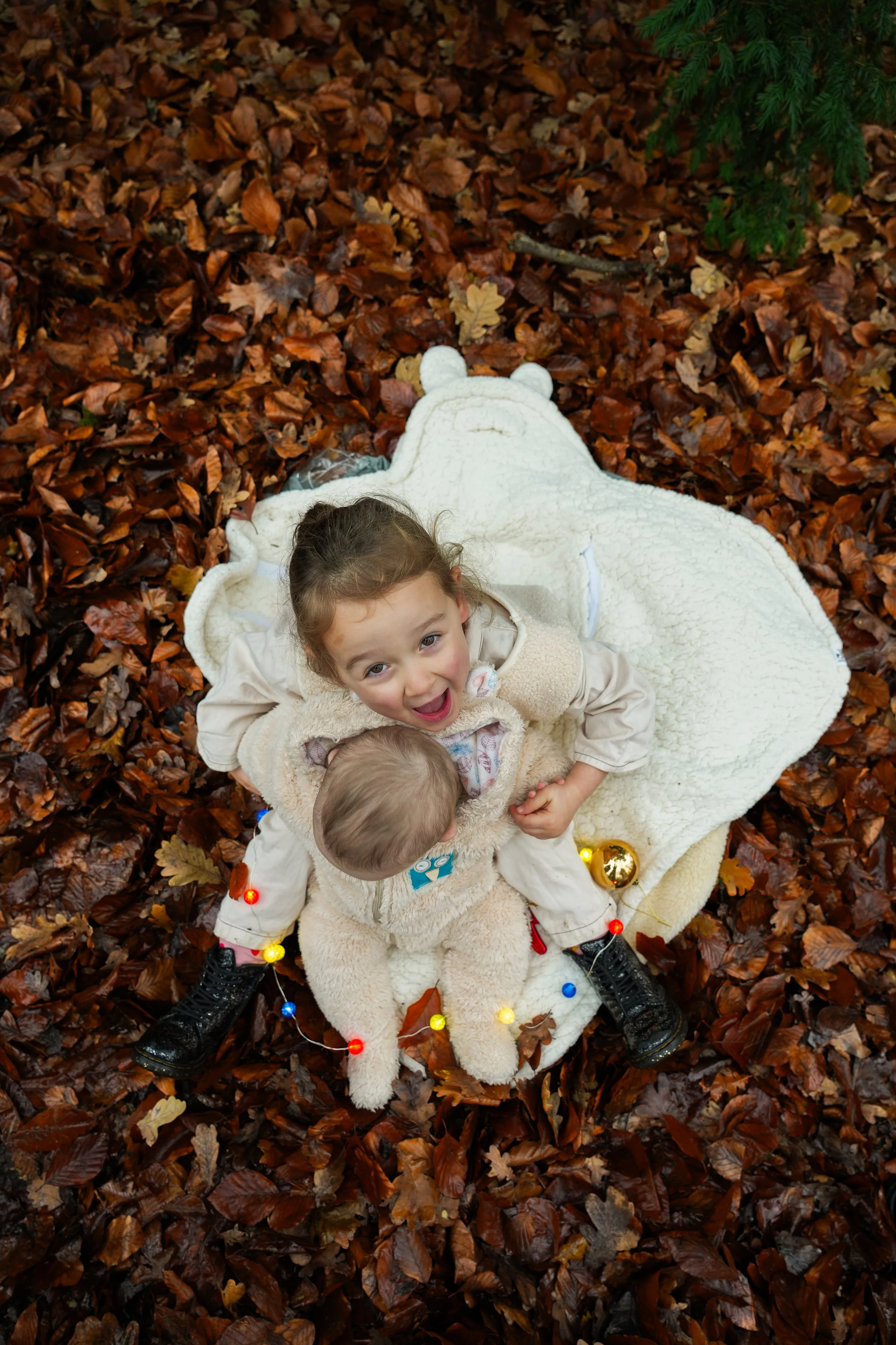 Two children, a girl and a baby, sit on a white blanket on fallen autumn leaves. The girl is smiling and looking up at the camera, holding the baby. The baby is dressed in a beige furry outfit. There are colorful Christmas lights around their feet an