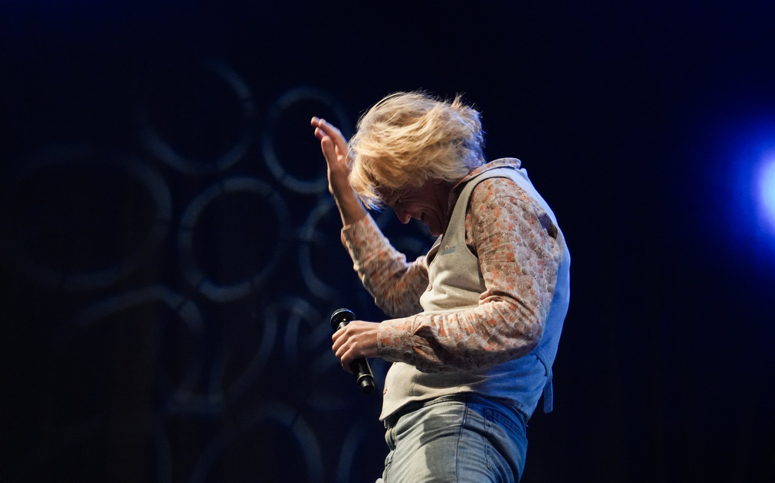 A woman with curly blonde hair on stage, holding a microphone in her right hand, laughing and raising her left hand, with circular patterns glowing in the dark background.