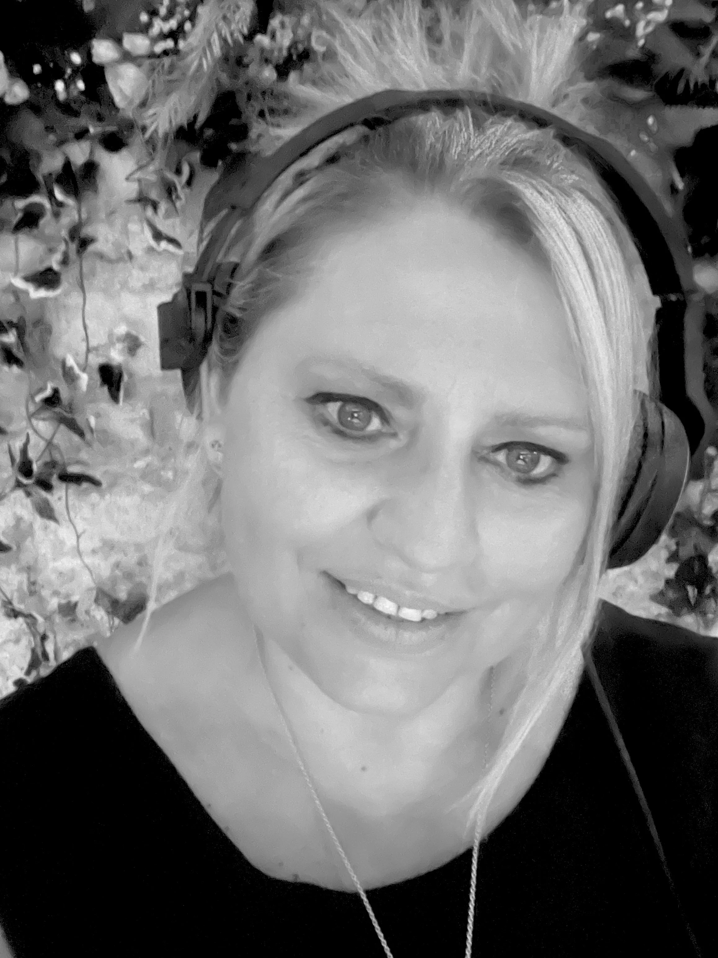 A photo of Claire smiling, wearing headphones around her neck and a black top, outdoors with leaves in the background.