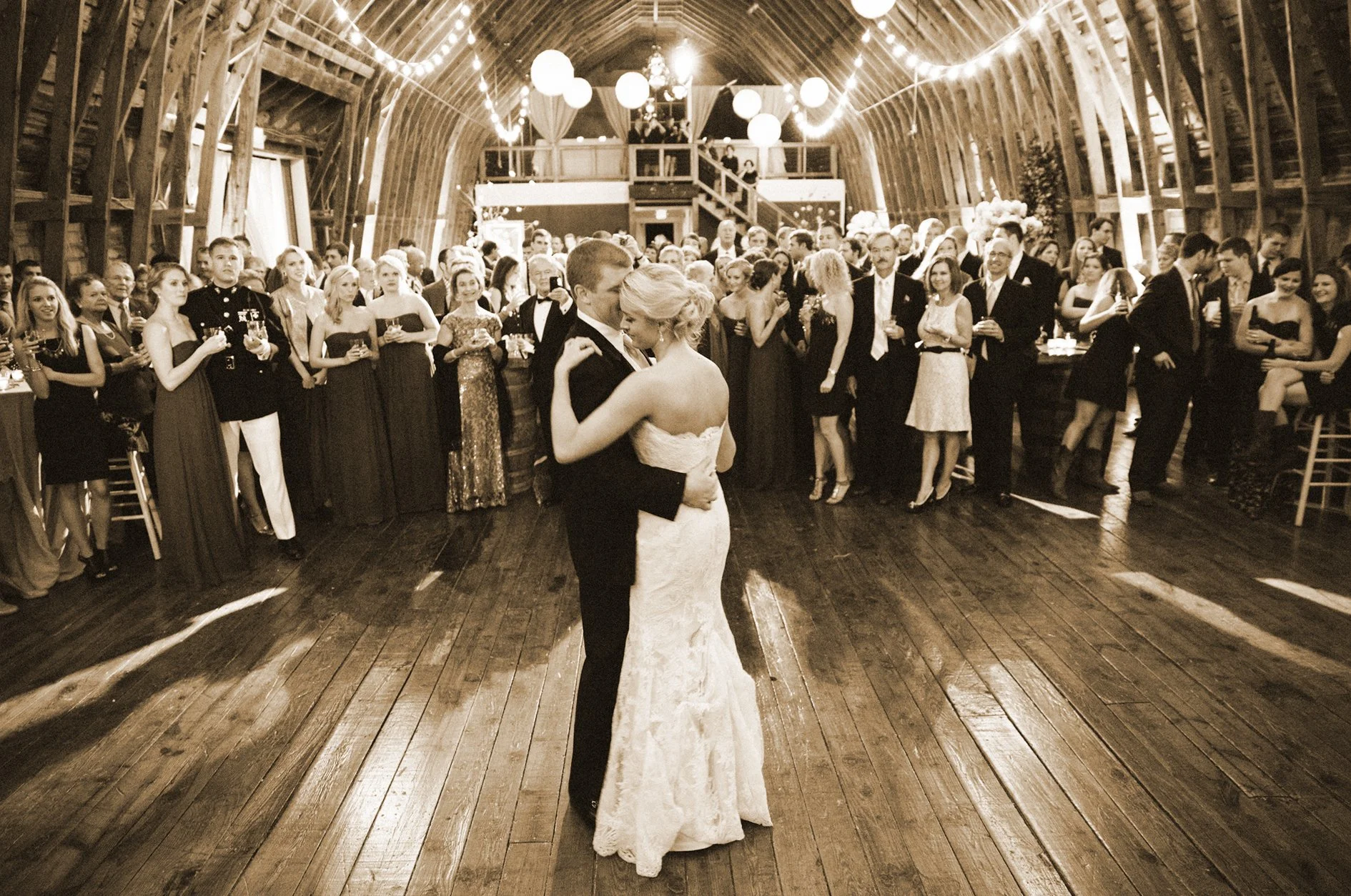 A bride and groom dancing in the center of a wooden dance floor at their wedding reception, surrounded by guests in an elegantly decorated barn-like venue.