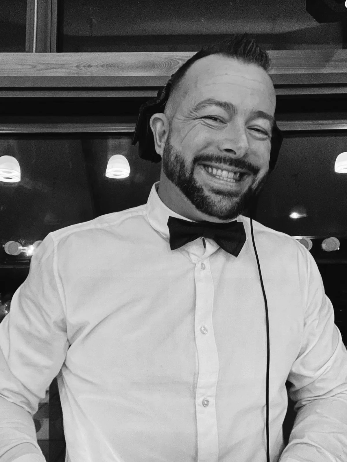 A man smiling, wearing a white shirt, black bow tie, and a headset, in an indoor setting with ambient lighting.