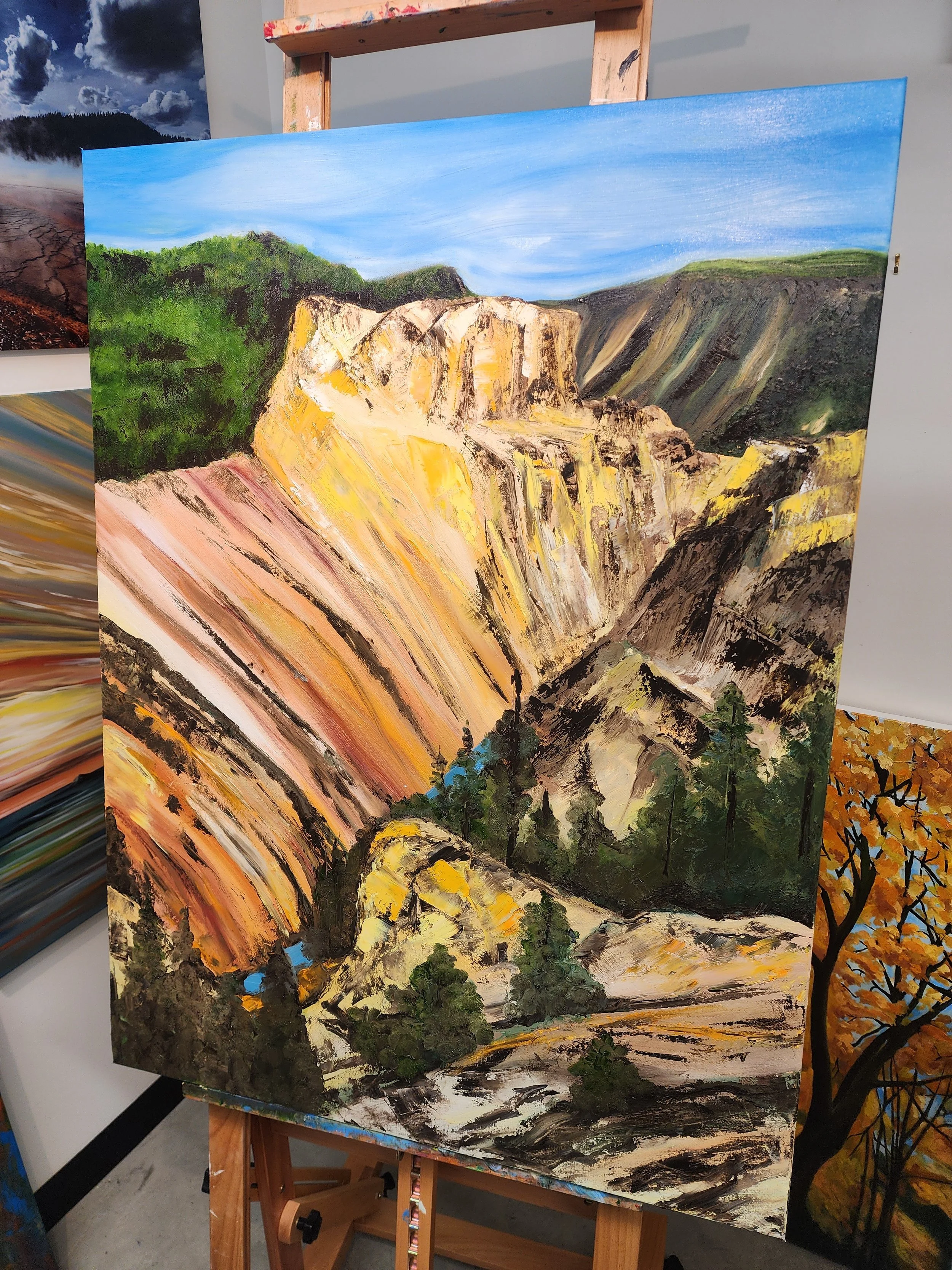 A landscape painting of a canyon with layered cliffs, green trees at the bottom, and a blue sky with wispy clouds.