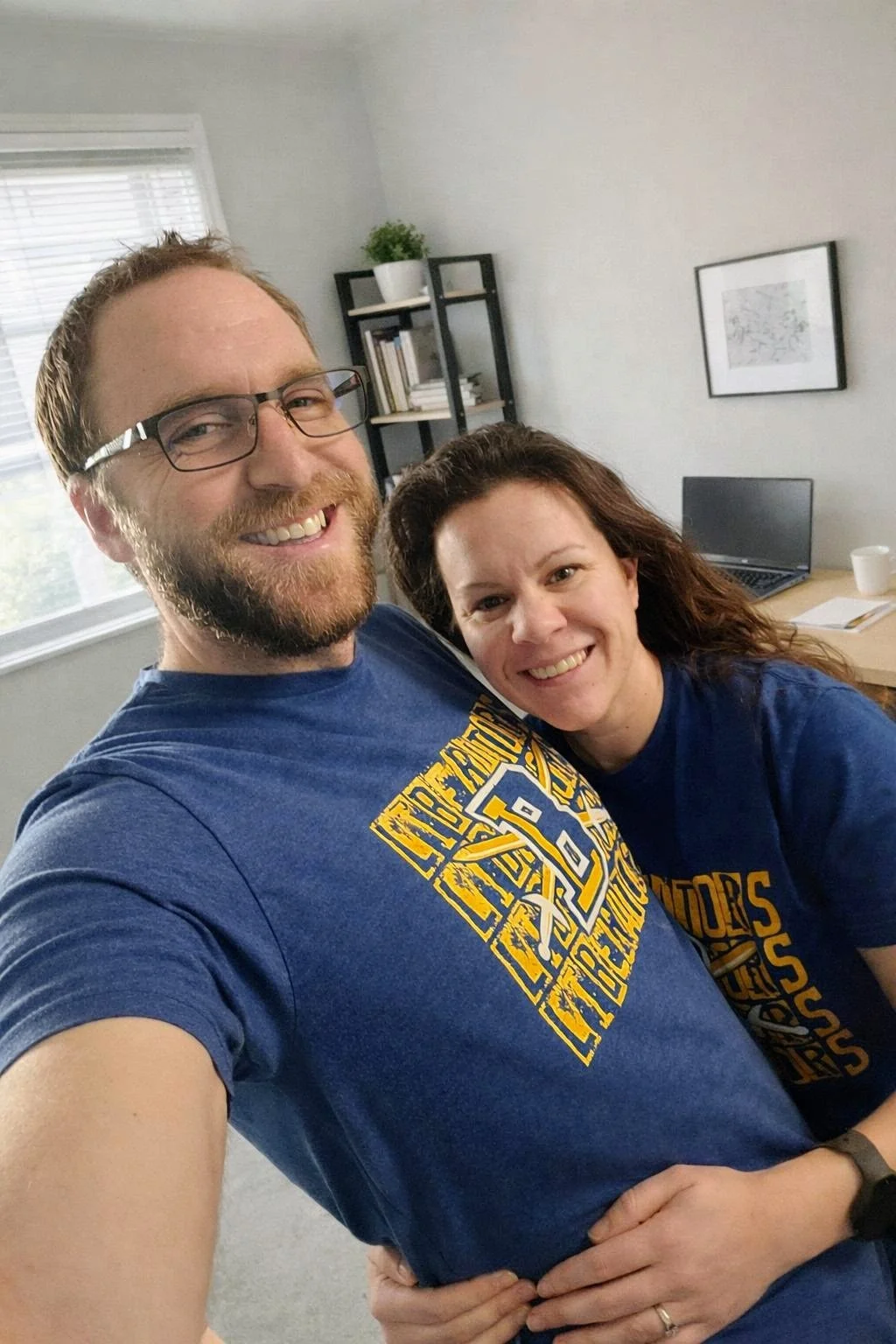 A man and a woman smiling and posing for a selfie in an office. The man has glasses and a beard, and the woman has long curly hair. They are both wearing blue shirts with yellow lettering.