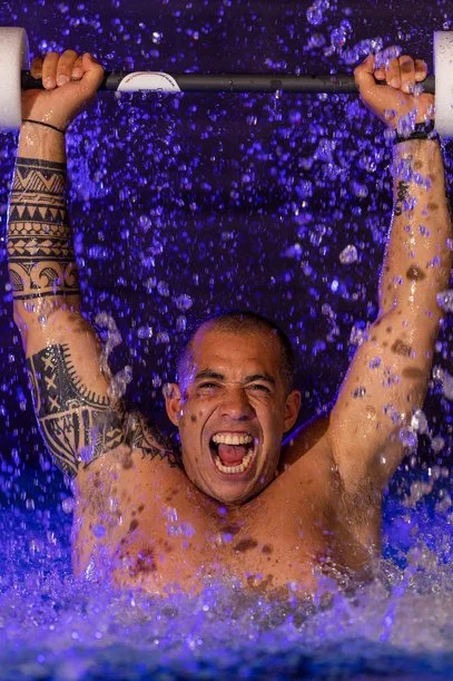 Een man die krachtig een halter boven zijn hoofd tilt in een waterbad, met water dat opspruit en een intense uitdrukking op zijn gezicht.