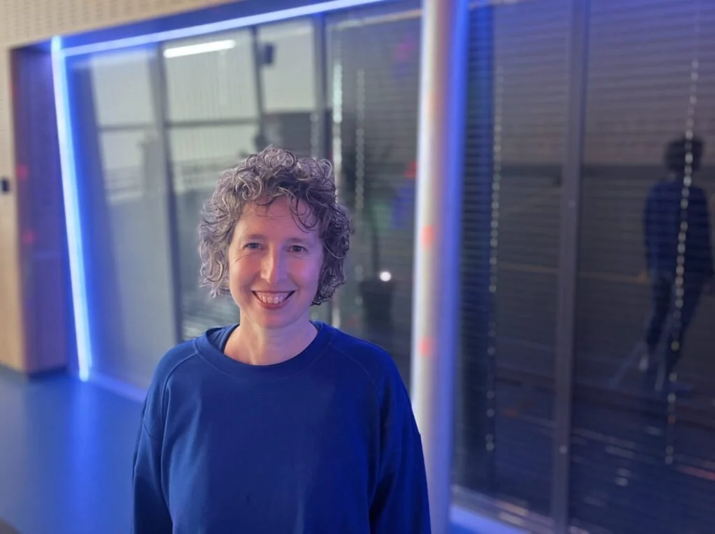 Een vrouw met gekruld haar glimlacht naar de camera, op de achtergrond neonverlichting en een glazen muur.