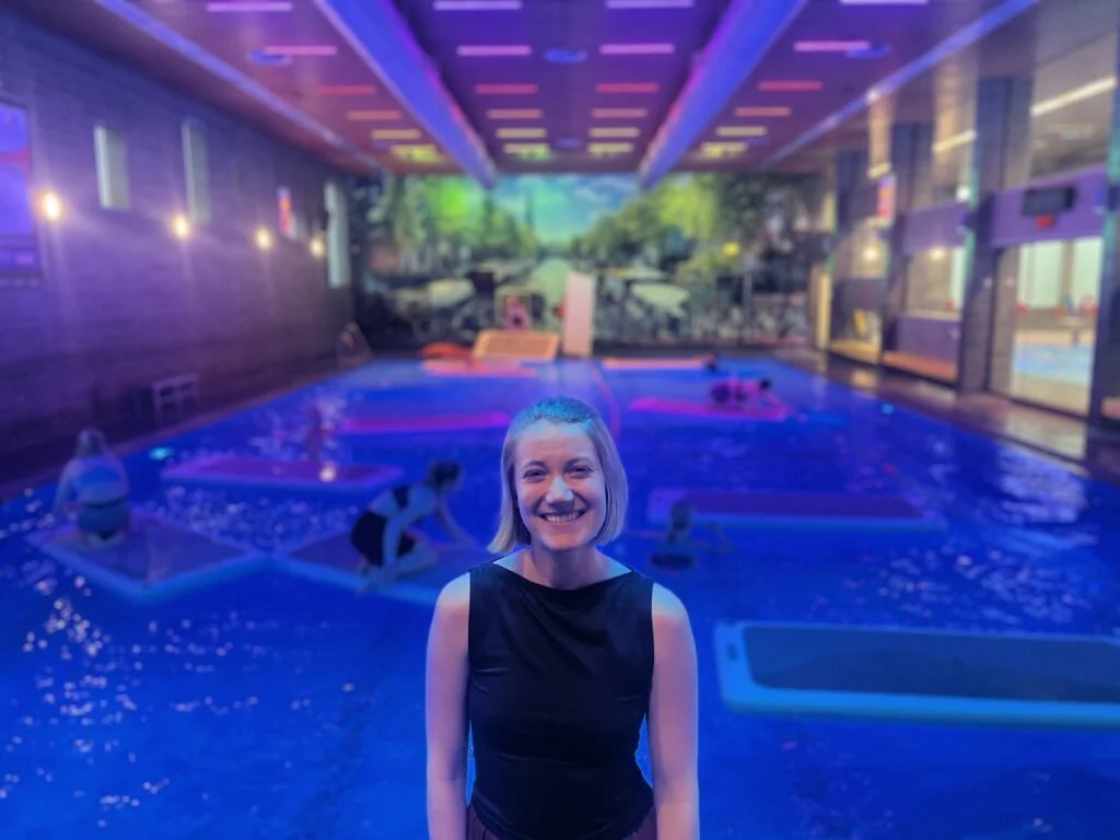 Vrouw in zwart topje glimlacht in een binnenzwembad met neonverlichting, met mensen die zwemmen en ontspannen op zwembadsutra