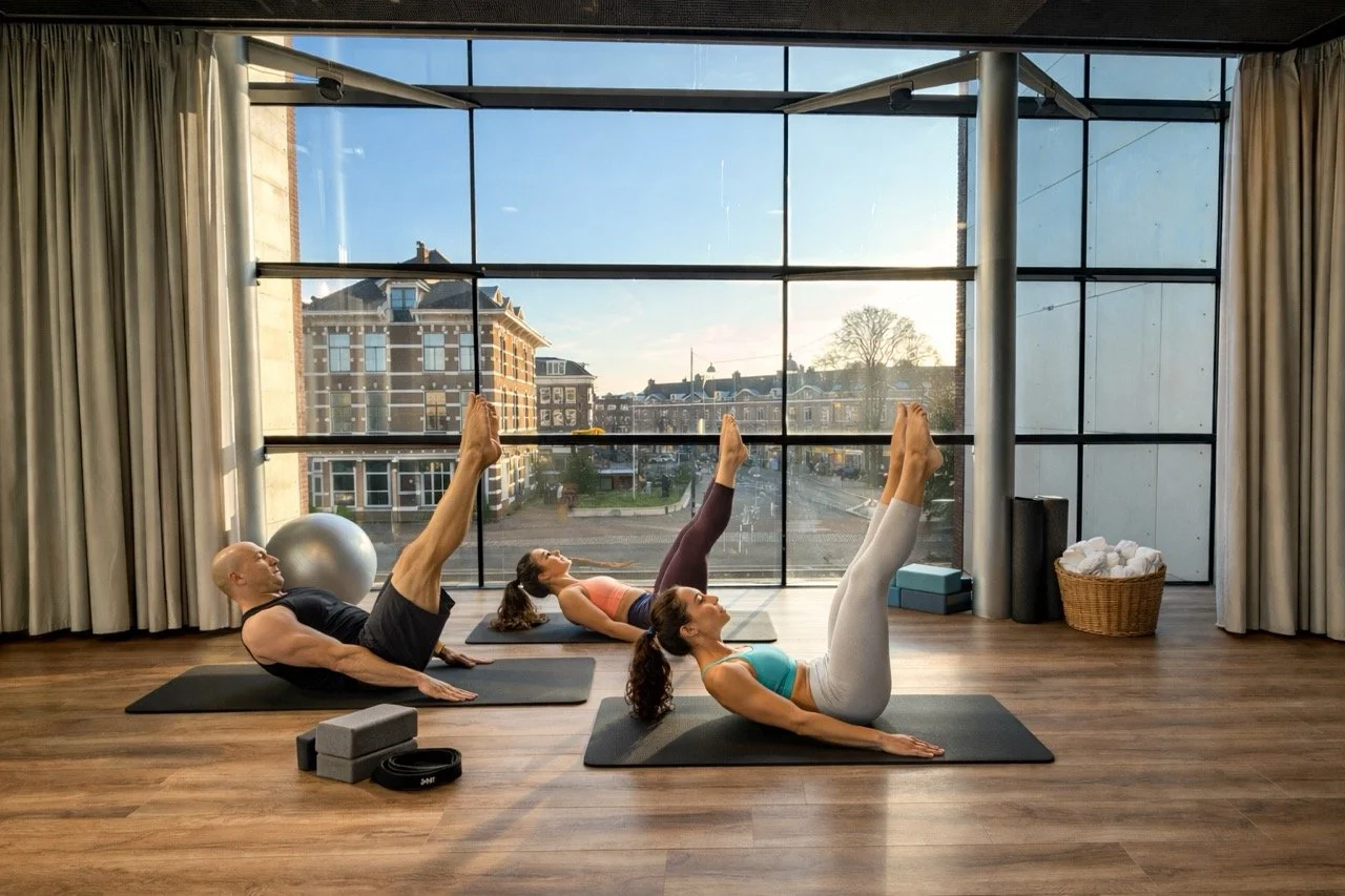 Drie mensen die yoga doen in een studio met grote ramen en een uitzicht op de stad.