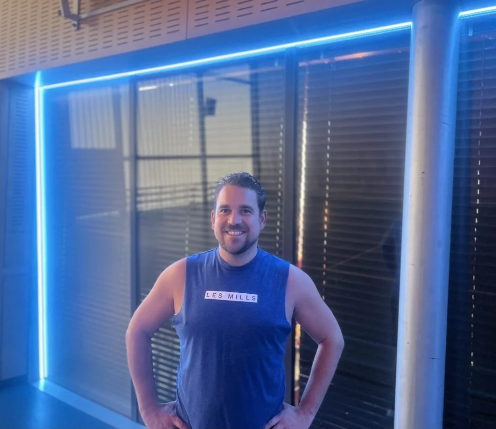 Man in een blauw sportshirt met tekst 'Les Mills' staat voor een moderne, verlichte muur met blauwe neonlichten.