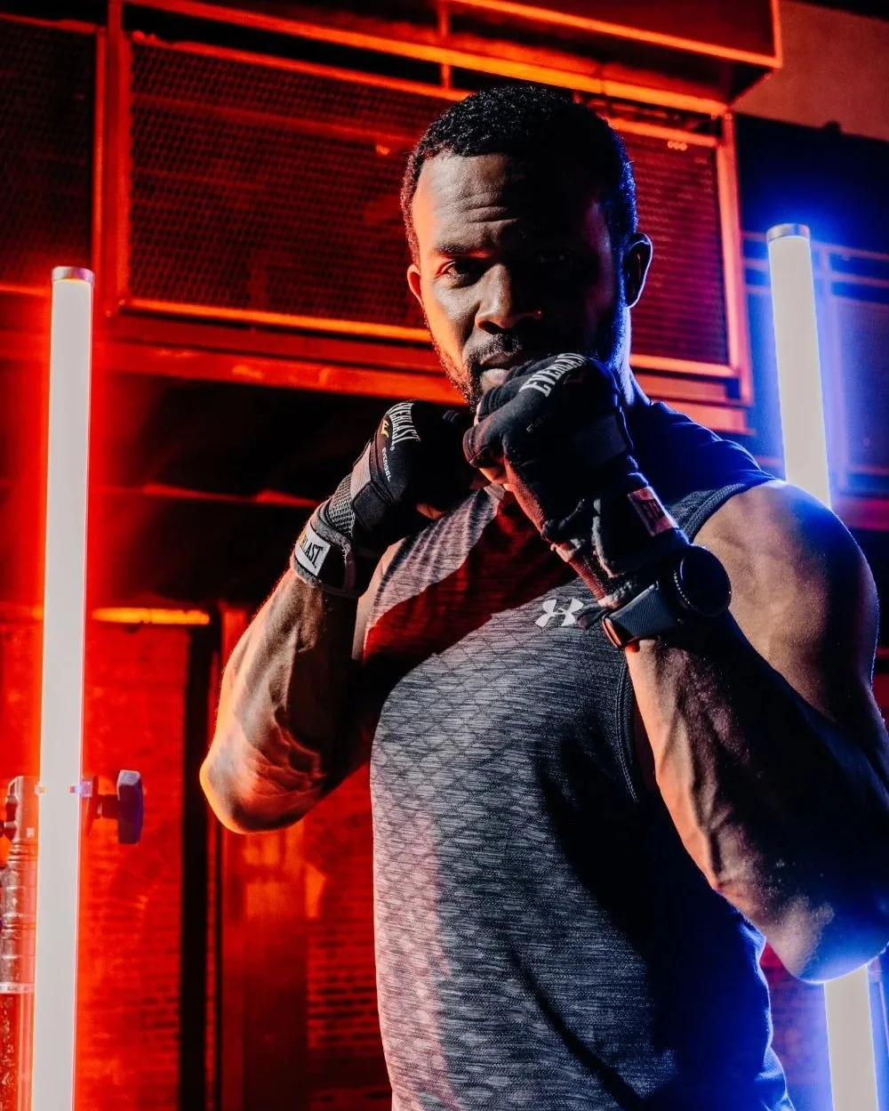 Een man in sportkleding en bokshandschoenen poseert in een sportschool met neonlichten.